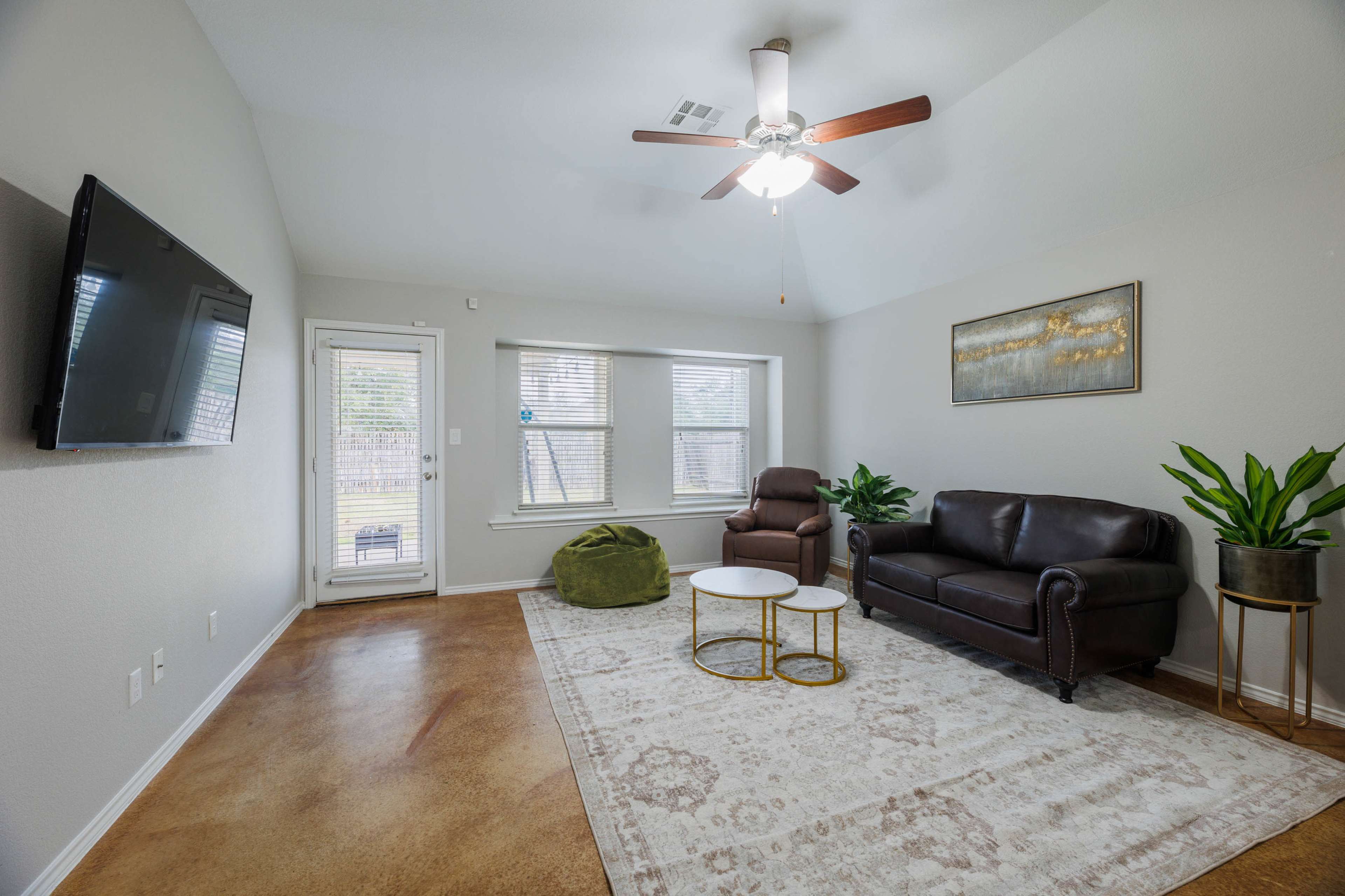 A living room features a leather couch, a recliner, a coffee table, and a ceiling fan, with large windows and a door leading outside.