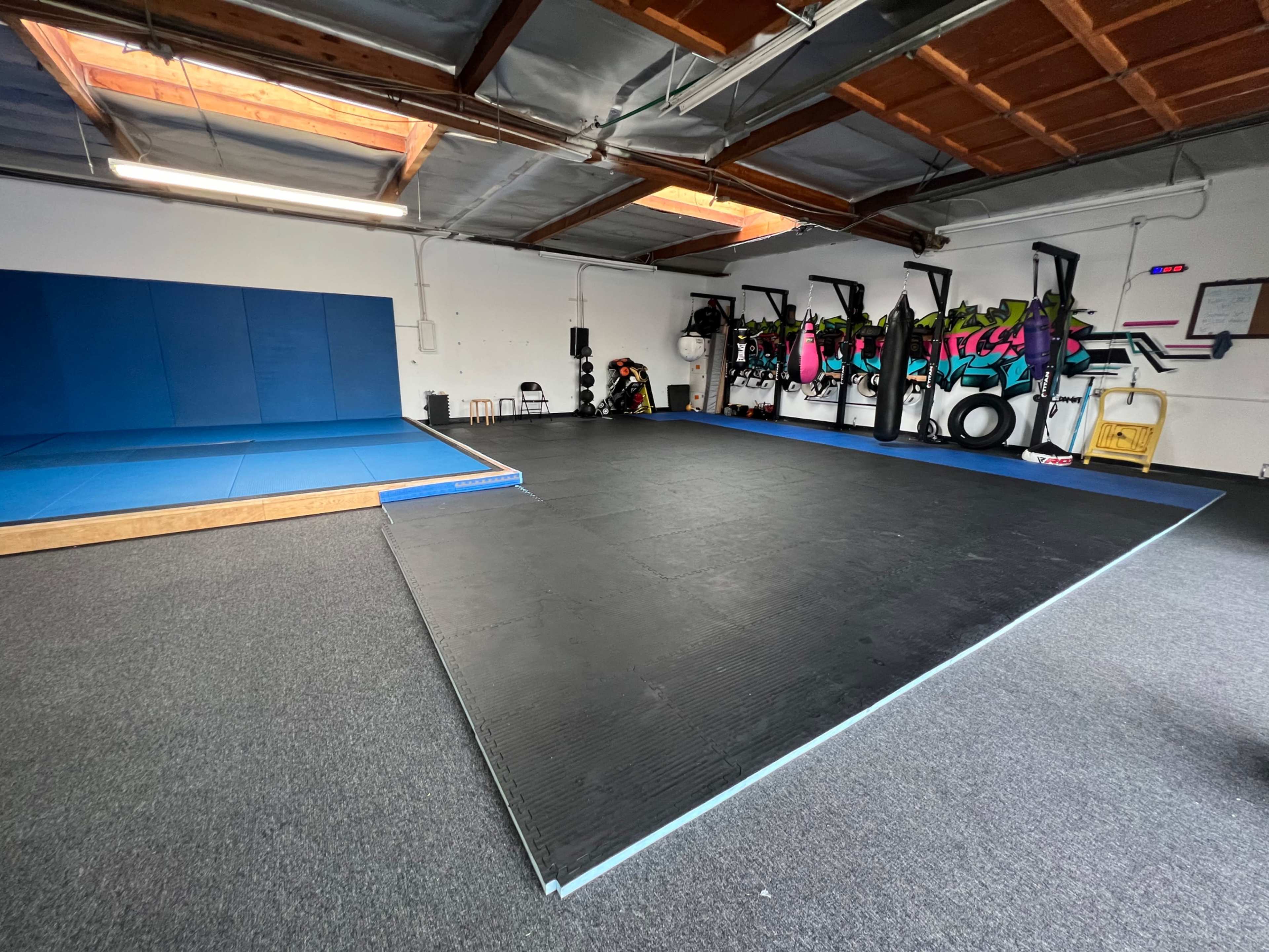 A training facility with a blue mat area and a black rubber floor, equipped with fitness equipment and wall decorations.