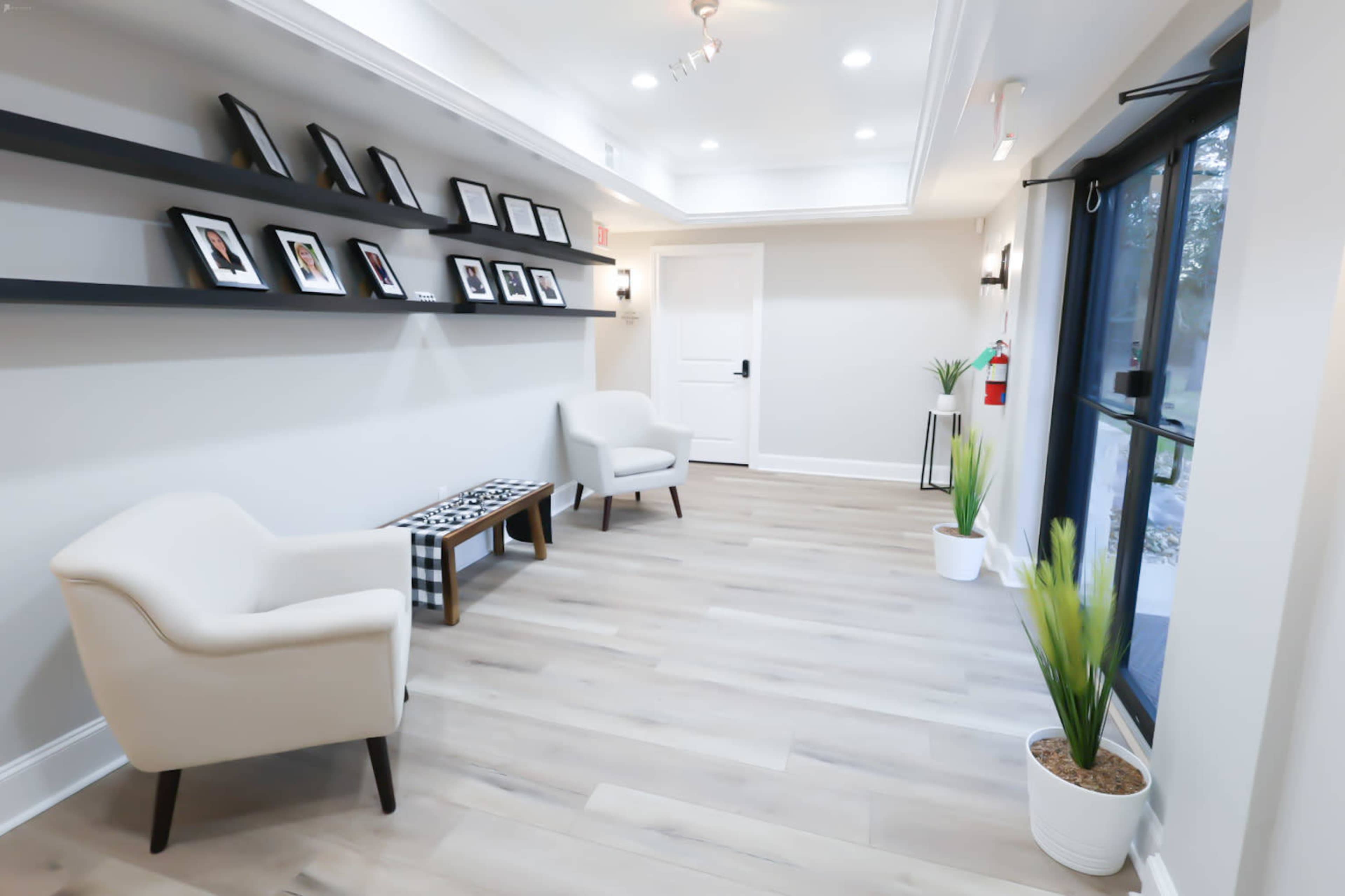 A bright hallway features two white chairs and a shelf displaying framed photographs along the wall.