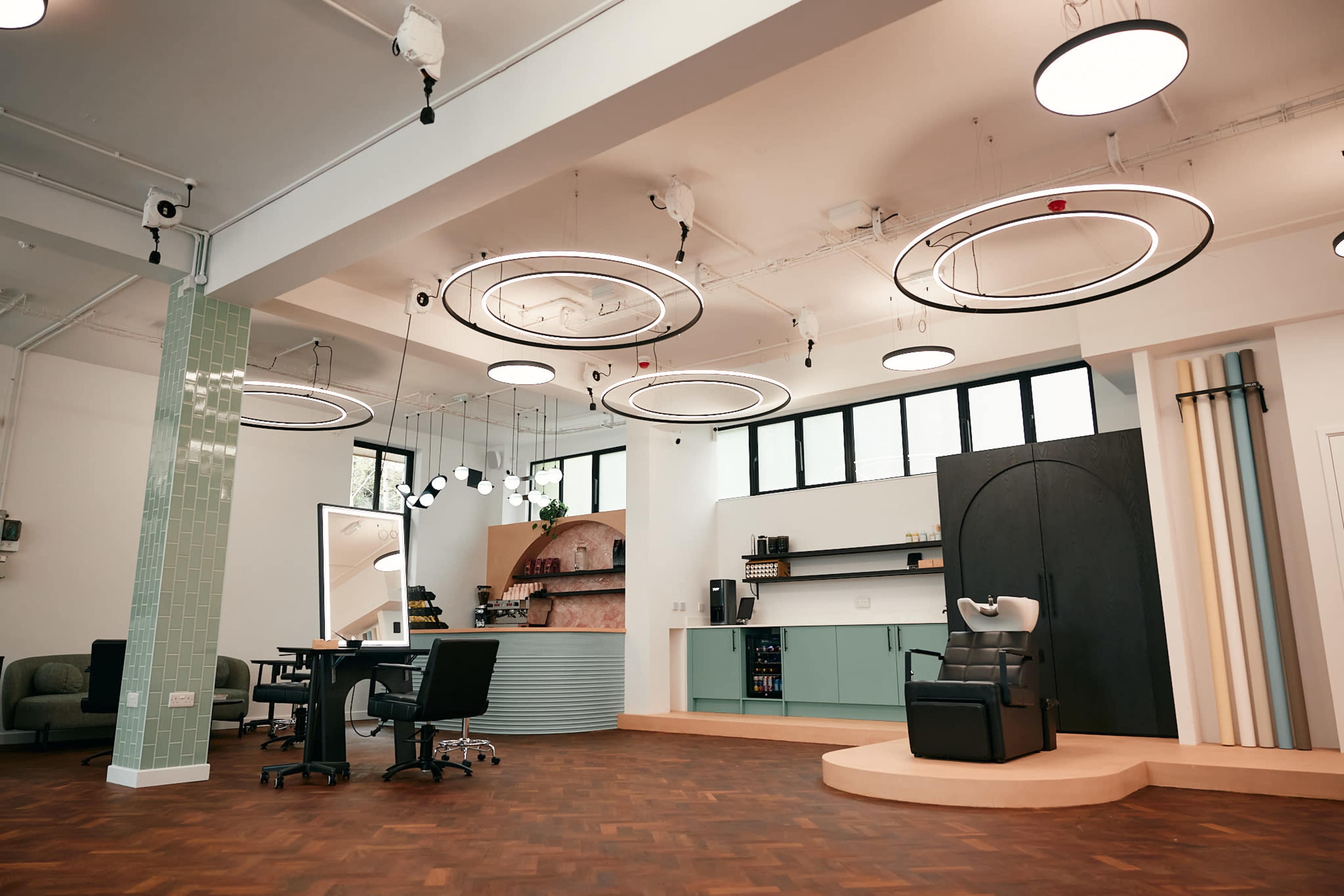 A modern hair salon with circular ceiling lights, a styling station, and a washing area, featuring a mix of green and neutral colors.