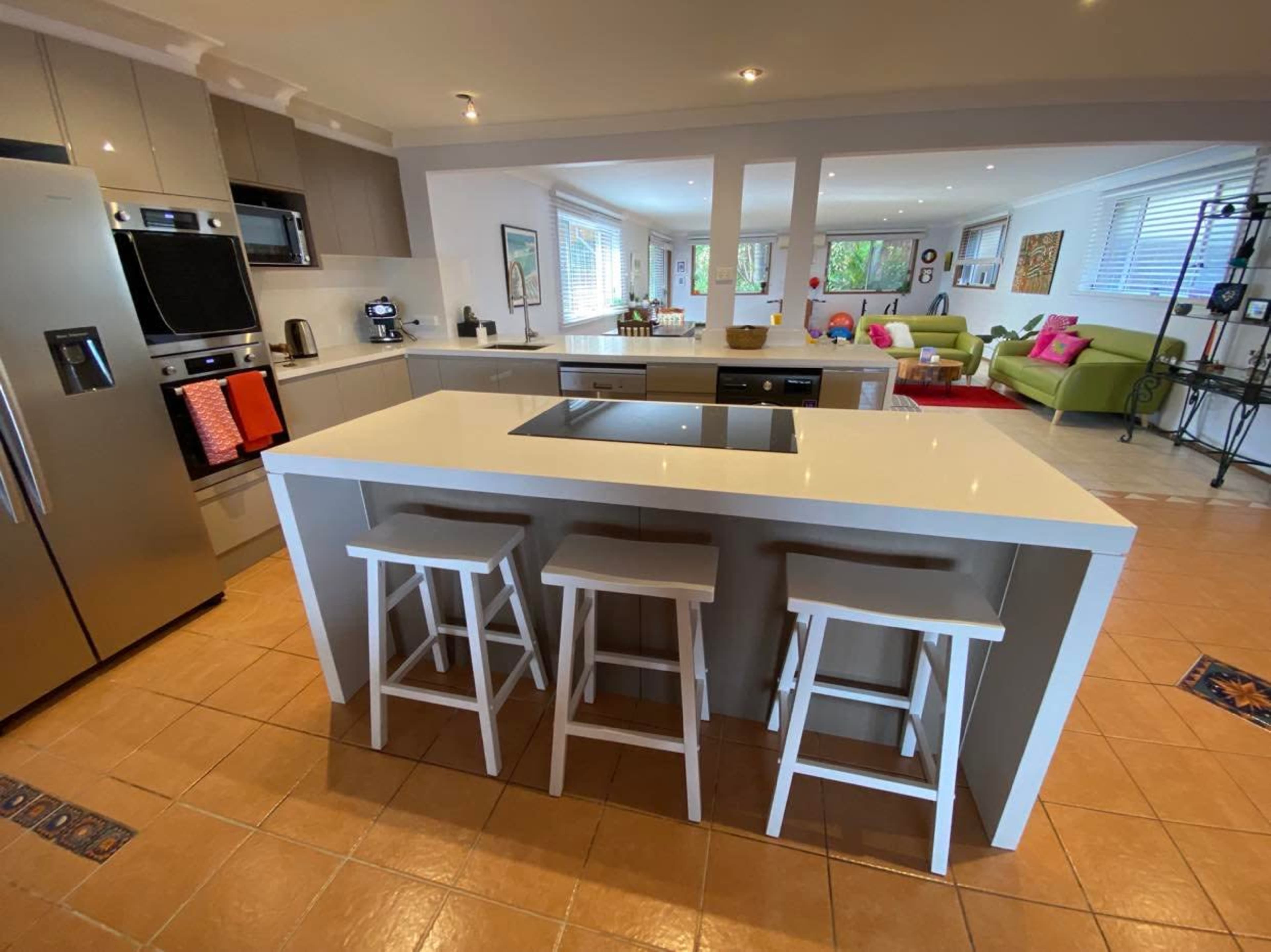 A modern kitchen with a central island, three white stools, stainless steel appliances, and an open layout leading to a living area.