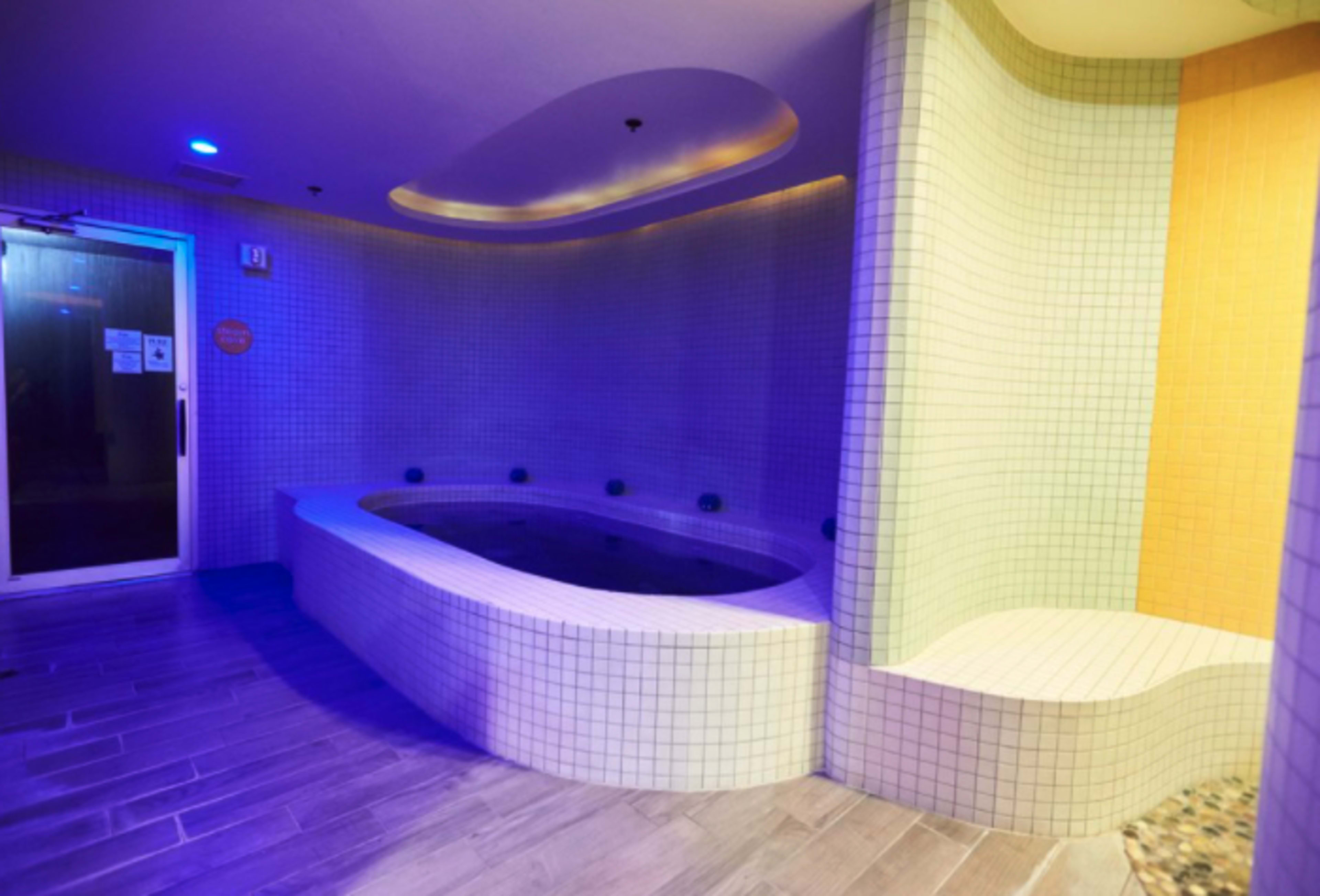 The image shows a tiled spa area with a curved seating area and a small hot tub, illuminated with soft blue lighting.