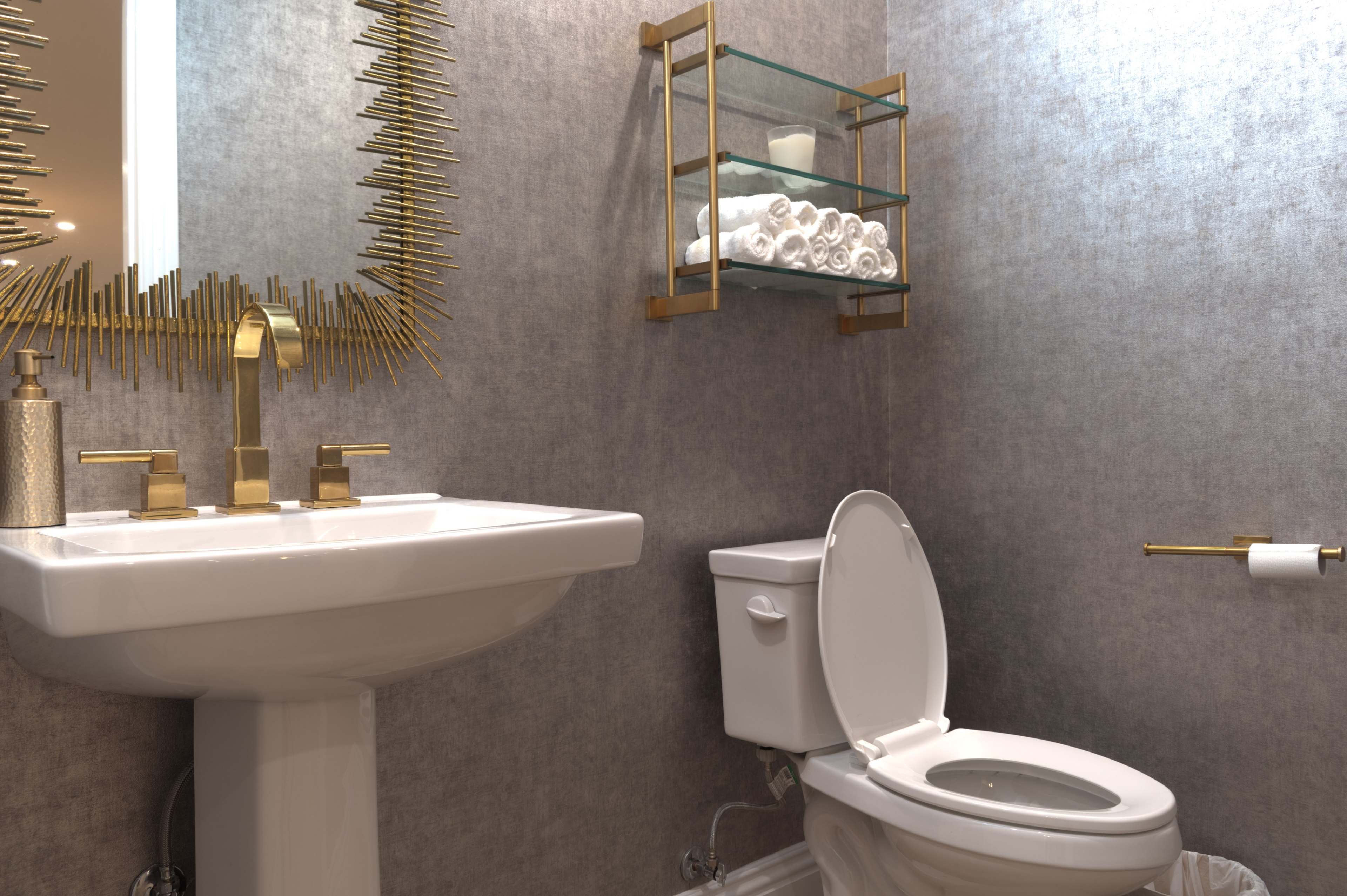 A modern bathroom featuring a pedestal sink, a toilet, a wall-mounted glass shelf with rolled towels, and a decorative mirror with a sunburst design.