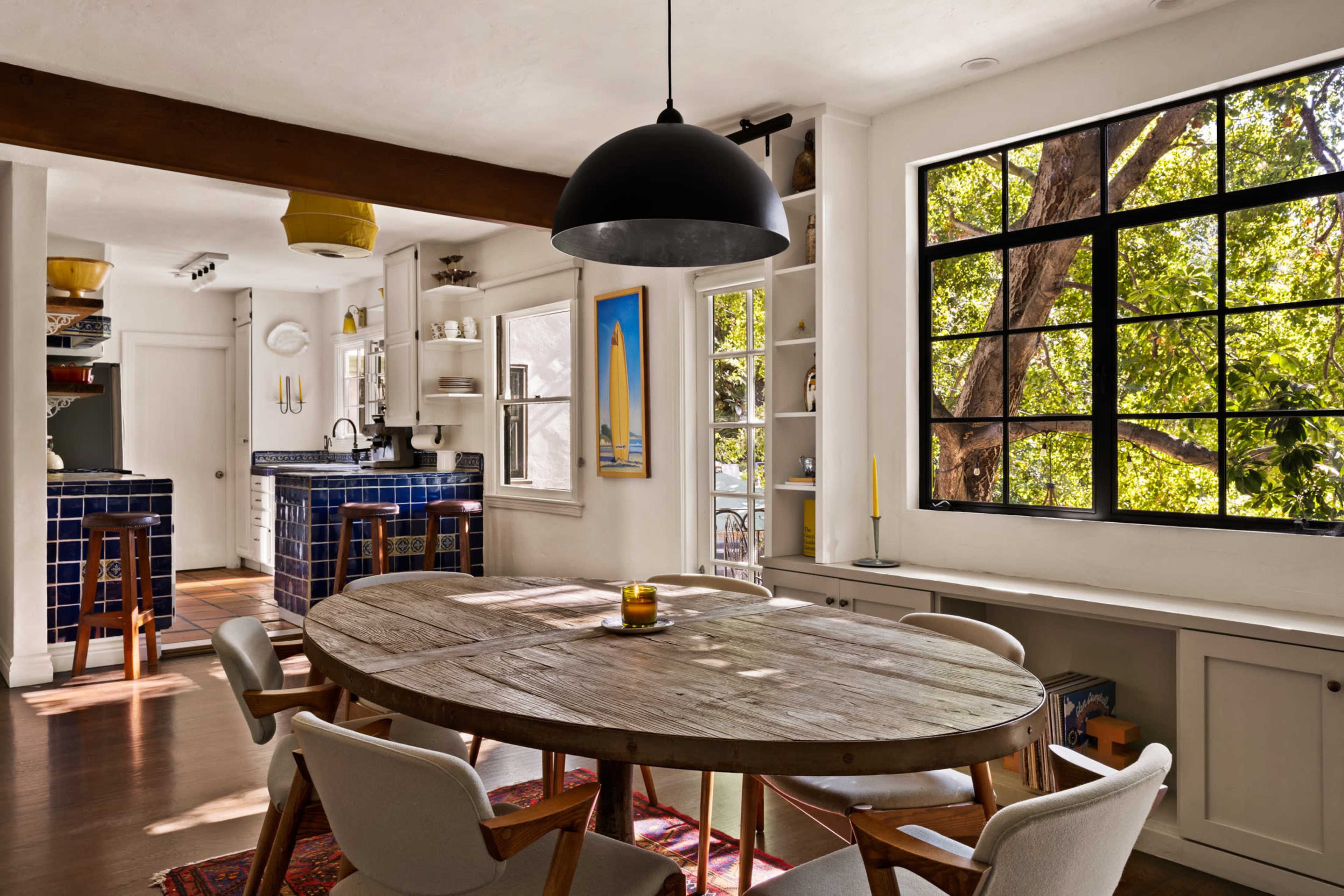 The image shows a bright kitchen and dining area featuring a round wooden table, modern chairs, and large windows that provide natural light and views of outdoor greenery.