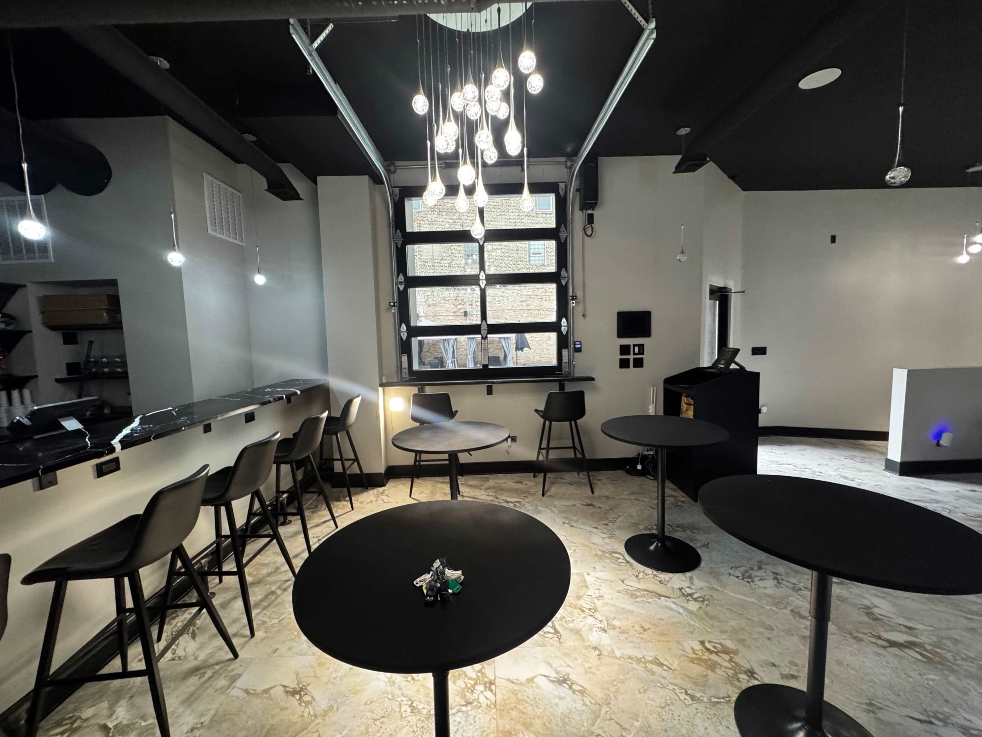 The image shows a modern interior of a bar or café, featuring high tables and stools, with large windows letting in natural light and a unique light fixture overhead.