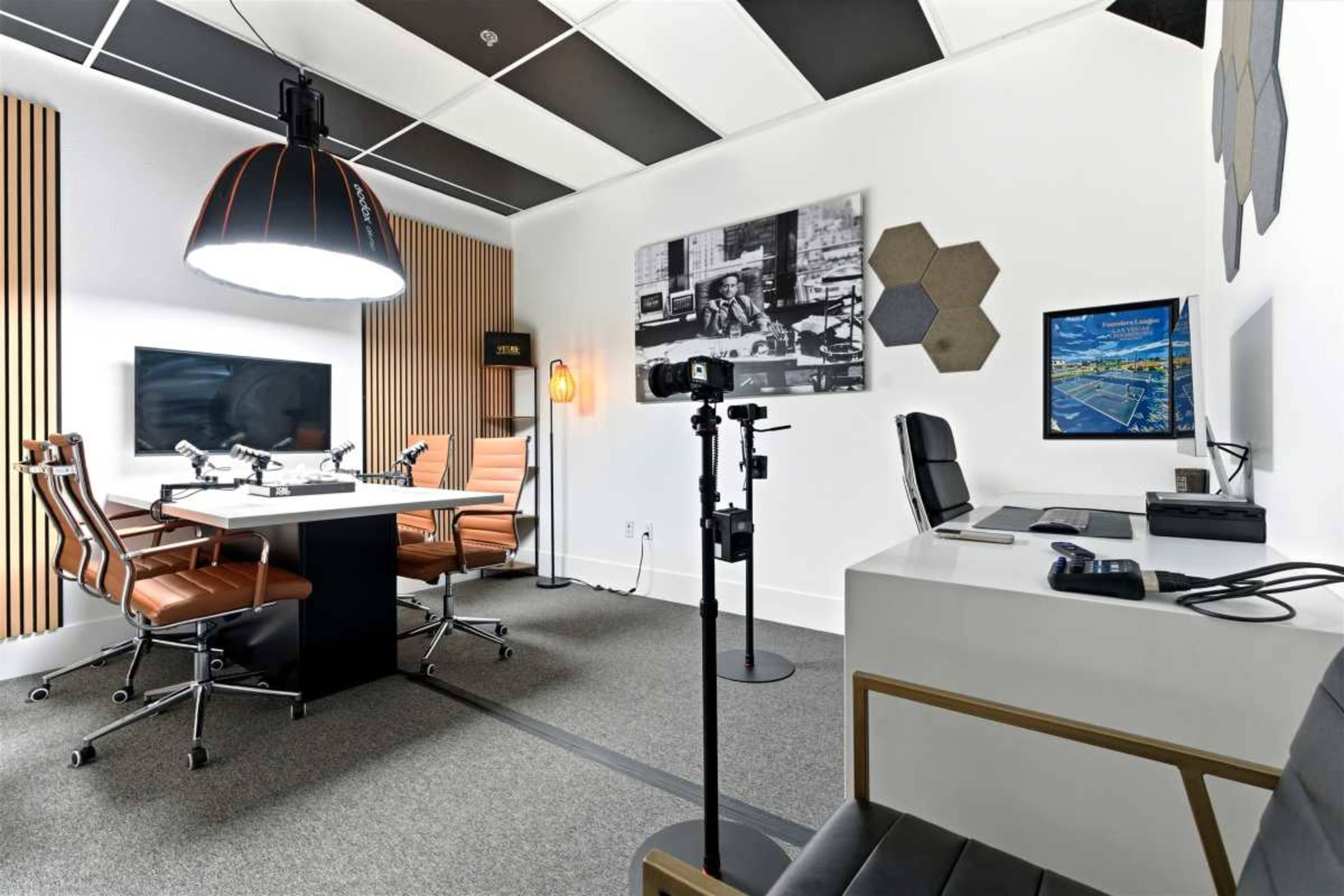 The image shows a modern office space featuring a conference table with leather chairs, a computer setup, and wall-mounted screens.
