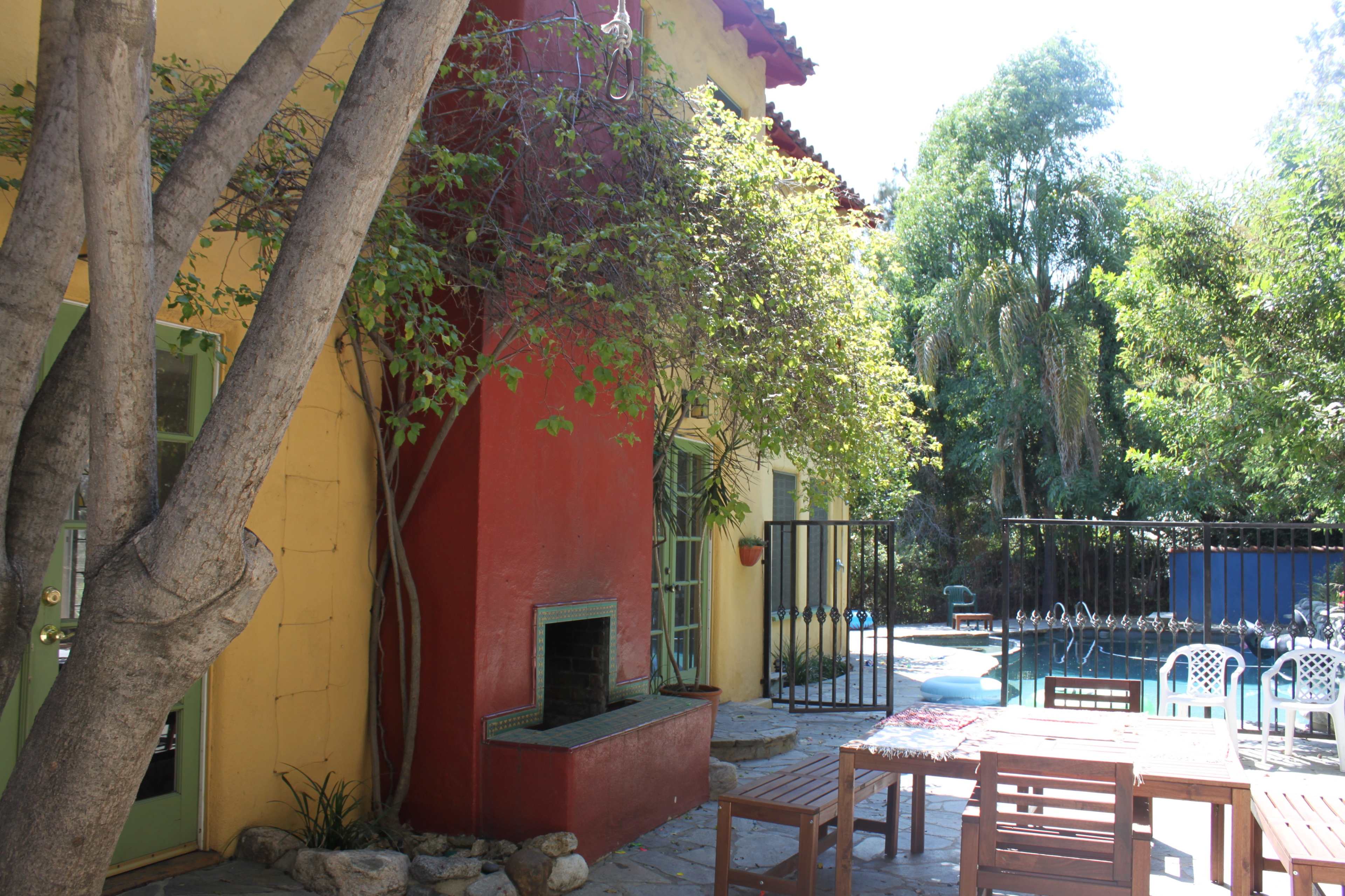 A terrace with wooden furniture is adjacent to a brightly colored house and a swimming pool surrounded by greenery.