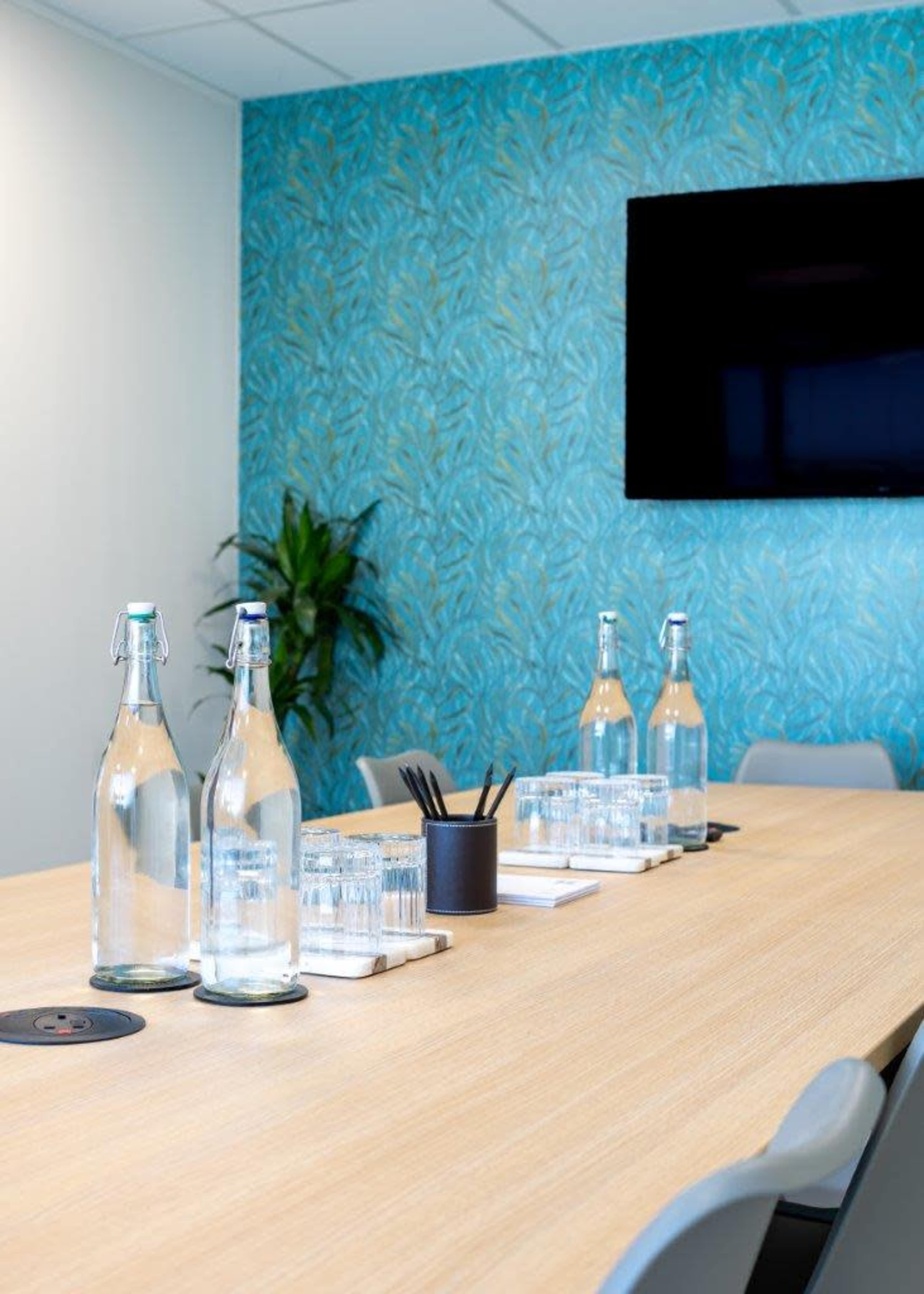 A modern conference room features a wooden table with several glass water bottles, glasses, and a black container for pens, set against a blue decorative wall and a mounted television.