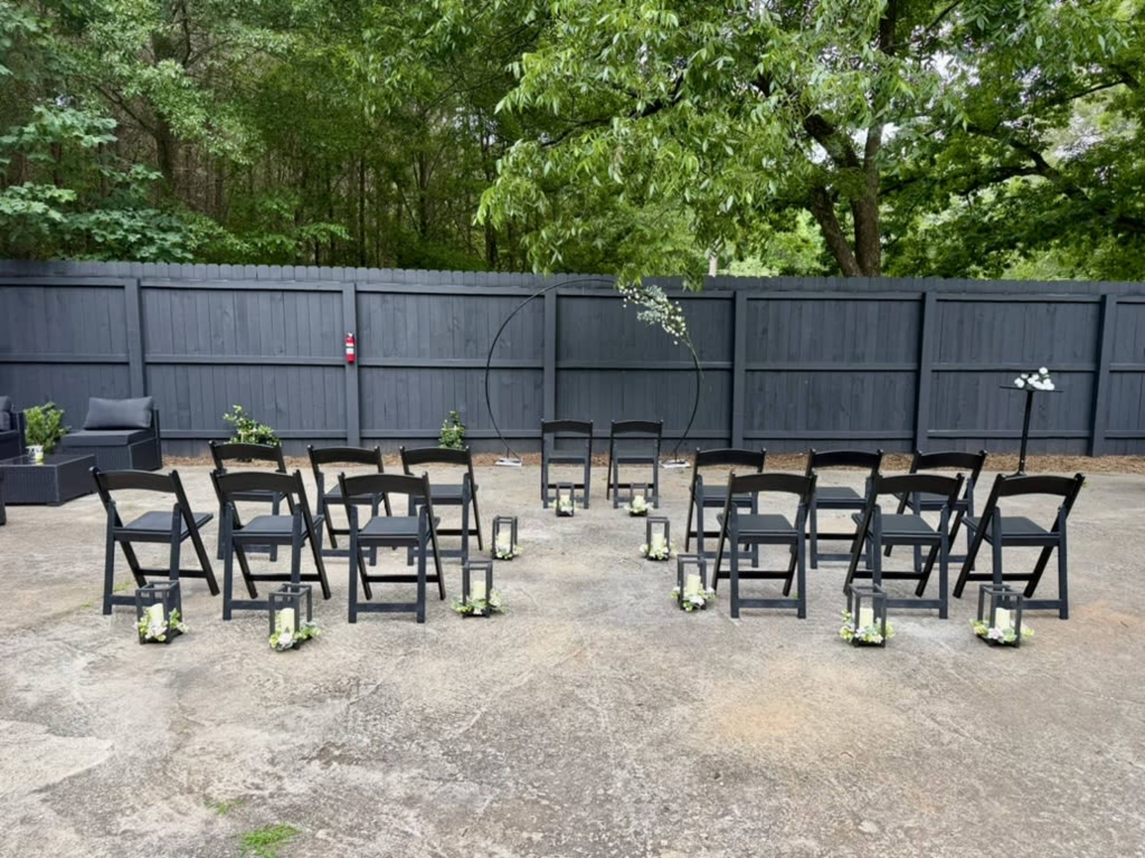 The image shows a decorated outdoor ceremony space with black folding chairs arranged in a semicircle around a circular arch, accented by lanterns and greenery.