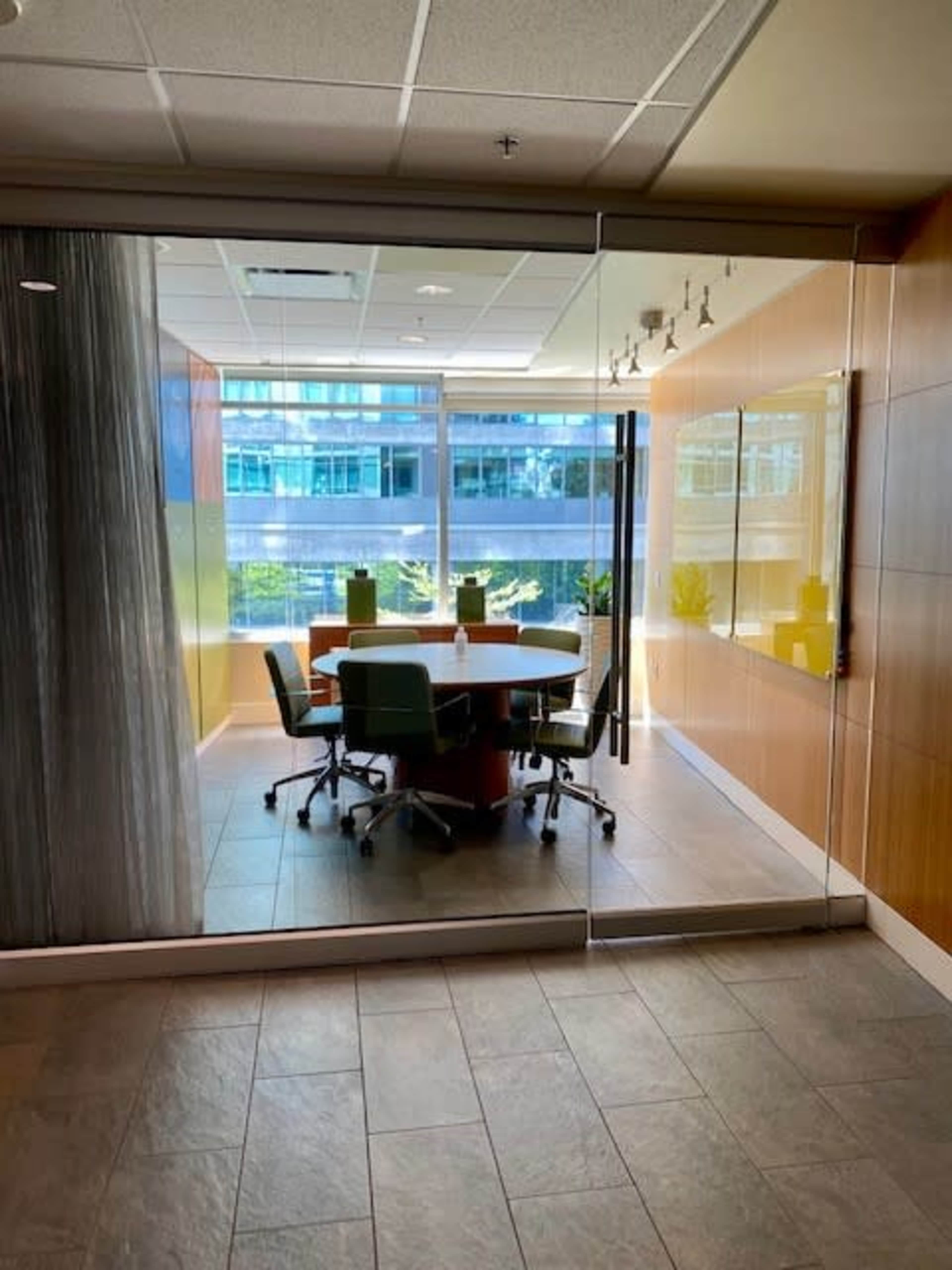 A glass-walled conference room features a round table surrounded by green chairs and large windows displaying the outside view.
