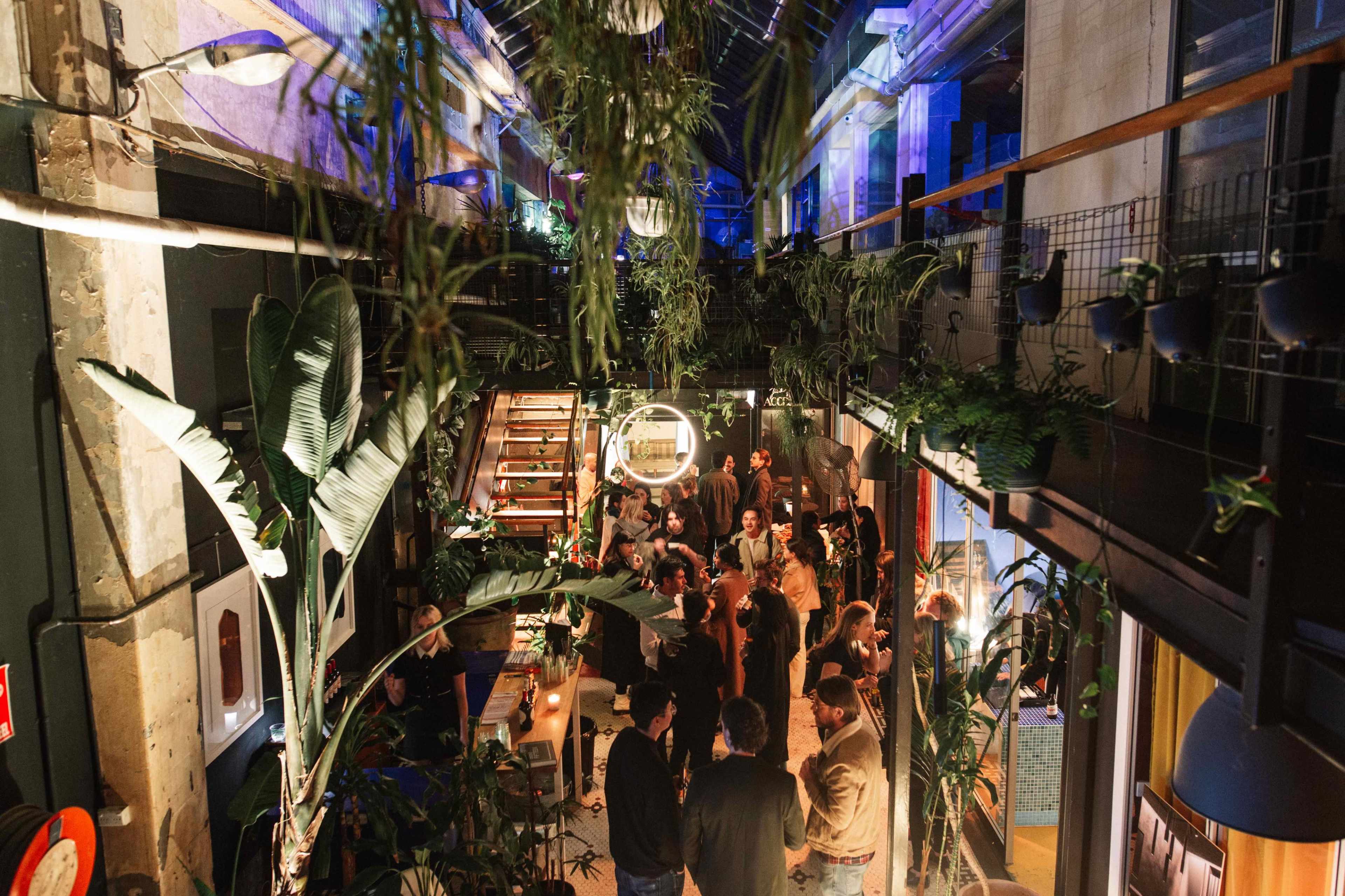 A vibrant gathering takes place in a spacious venue adorned with plants and soft lighting, as people socialize and mingle on multiple levels.