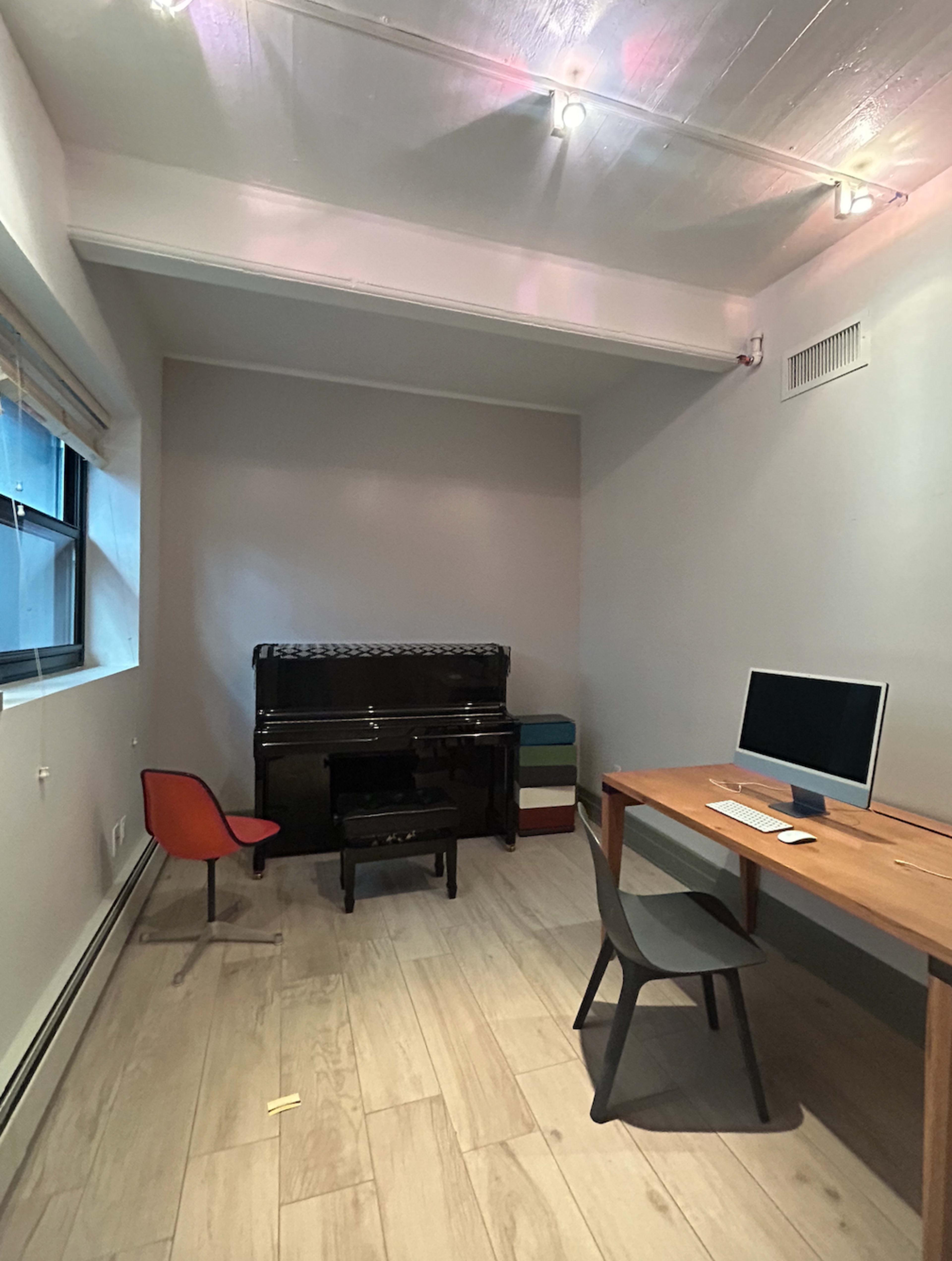 A minimal office space with a black piano, a desk with a computer, and two chairs, all set against light gray walls and wooden flooring.