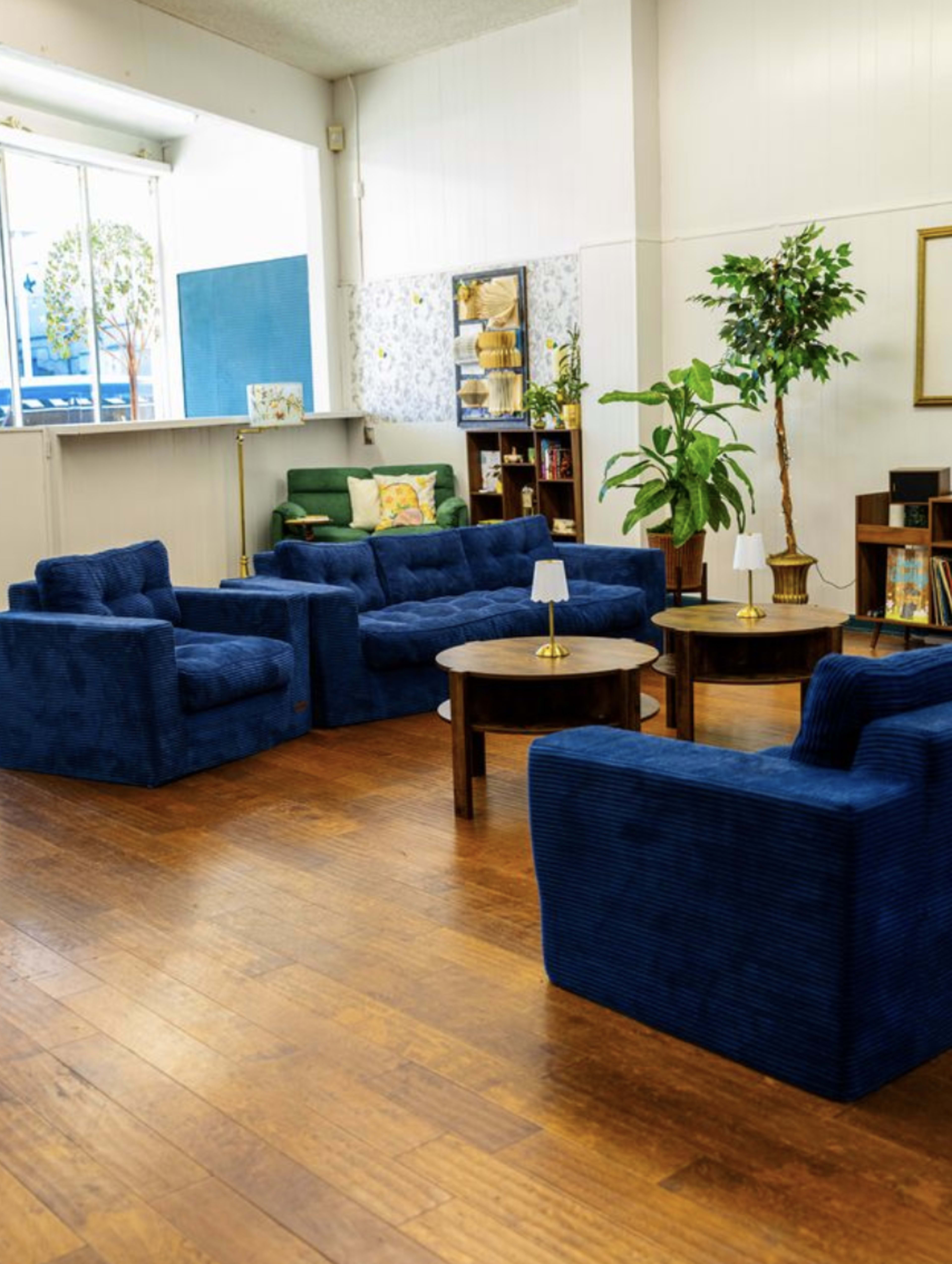 The image shows a cozy, furnished lounge area with blue armchairs and a wooden table, featuring plants and decorative elements on the walls.