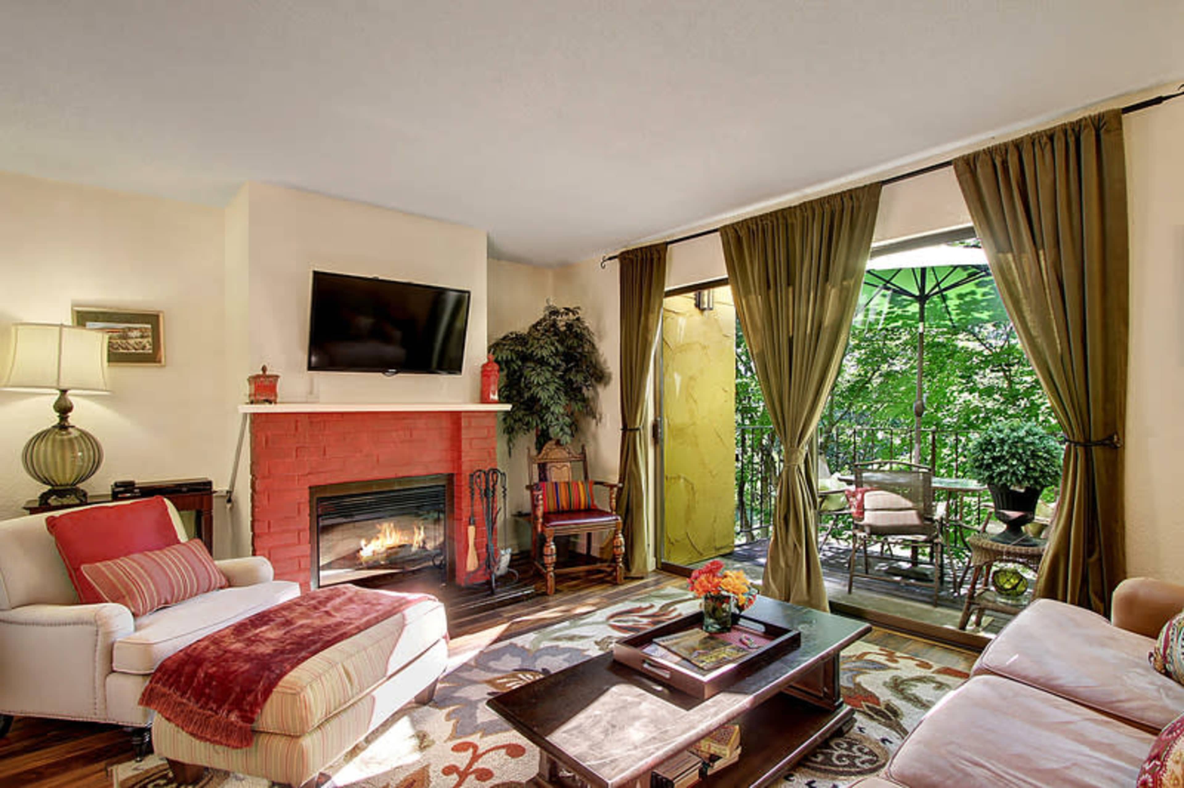 A cozy living room features a brick fireplace, a mounted television, and a view of a patio with greenery through large windows.