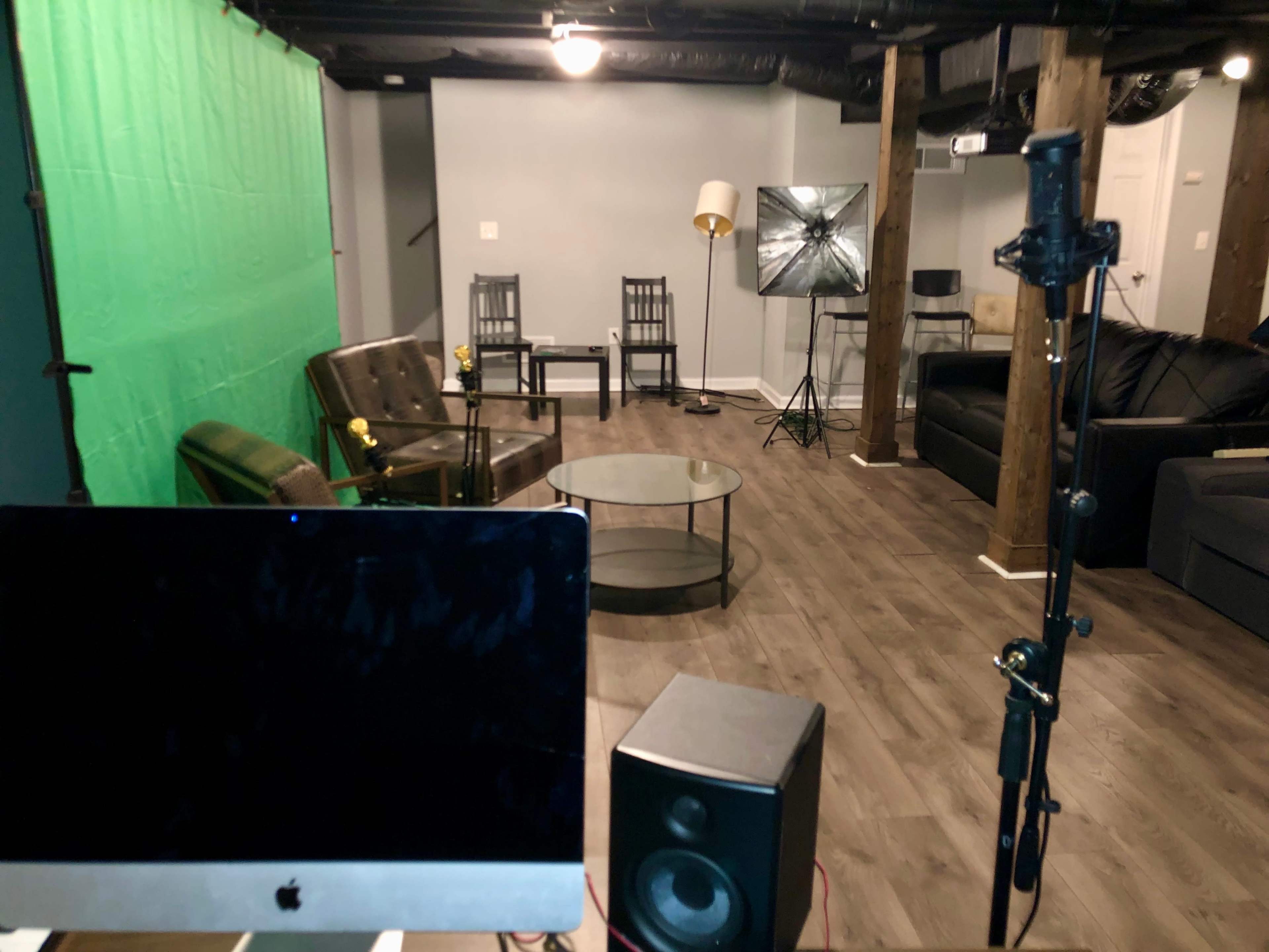 A basement studio setup featuring a green screen backdrop, two armchairs, a glass coffee table, a lamp, and various studio equipment.