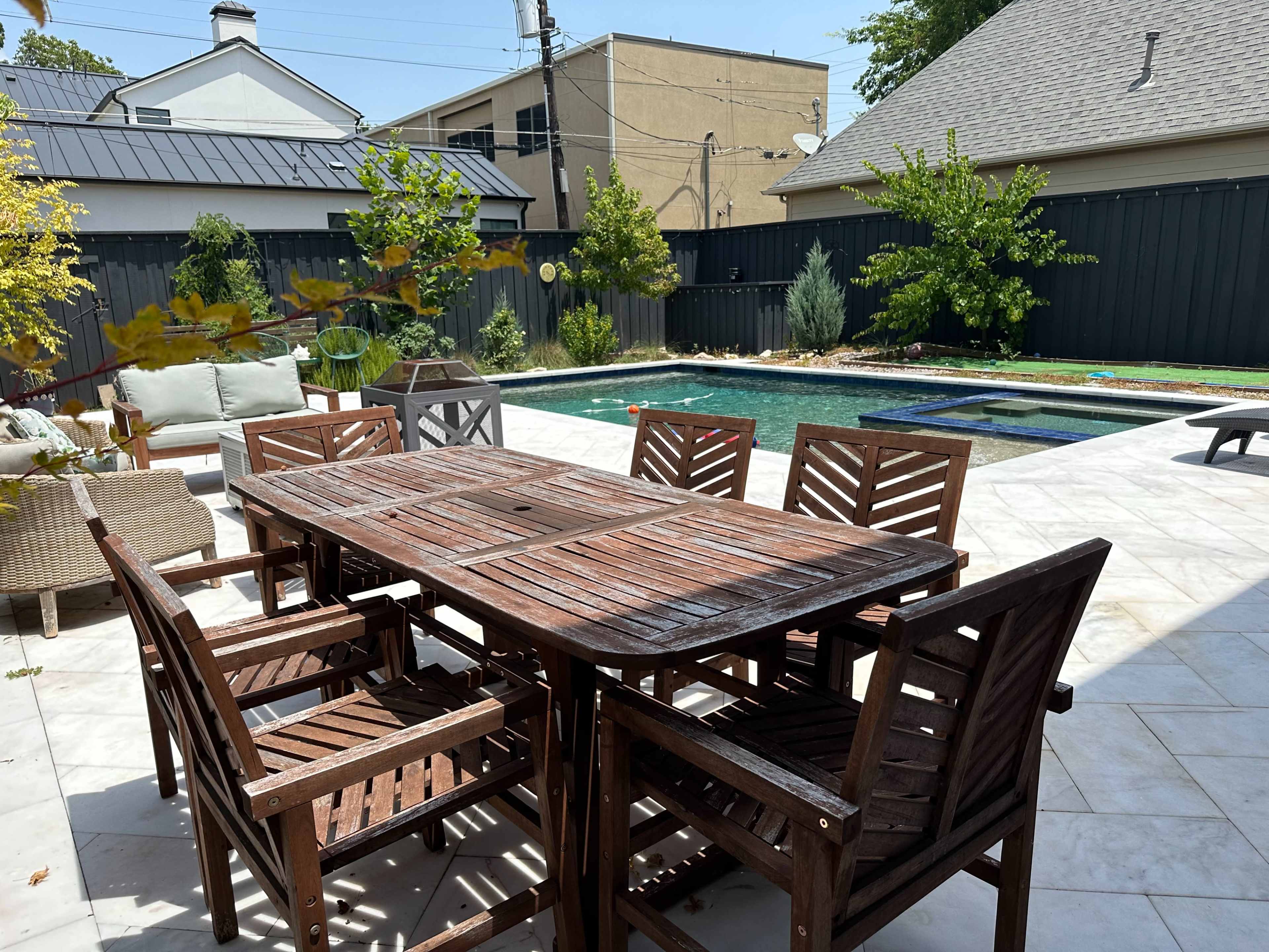 A wooden dining table surrounded by six chairs is set on a tiled patio near a swimming pool, with greenery and a fence in the background.