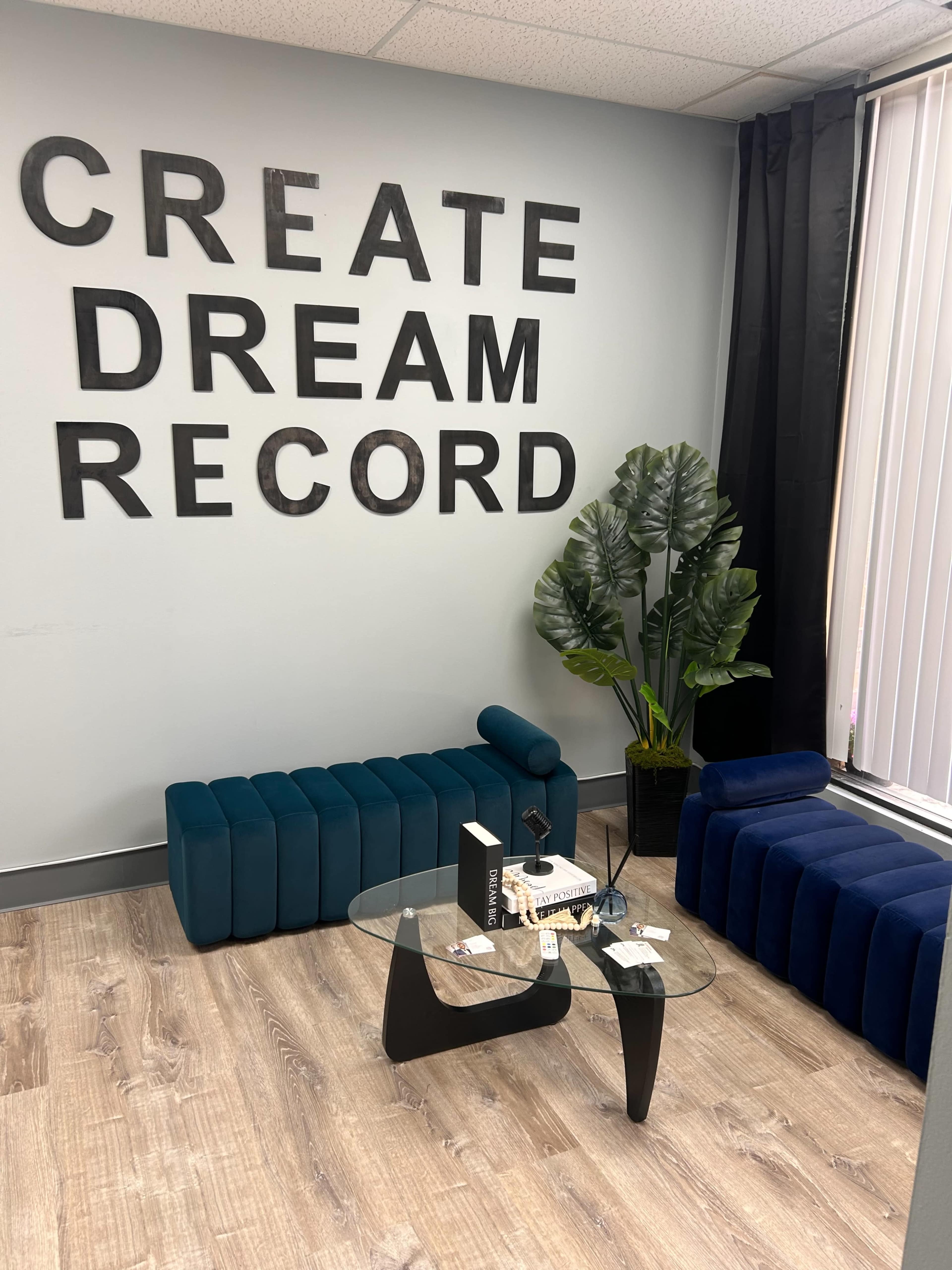 The image shows a modern lounge area with two teal seating sections, a glass coffee table, and a decorative plant against a wall featuring the words "CREATE DREAM RECORD."