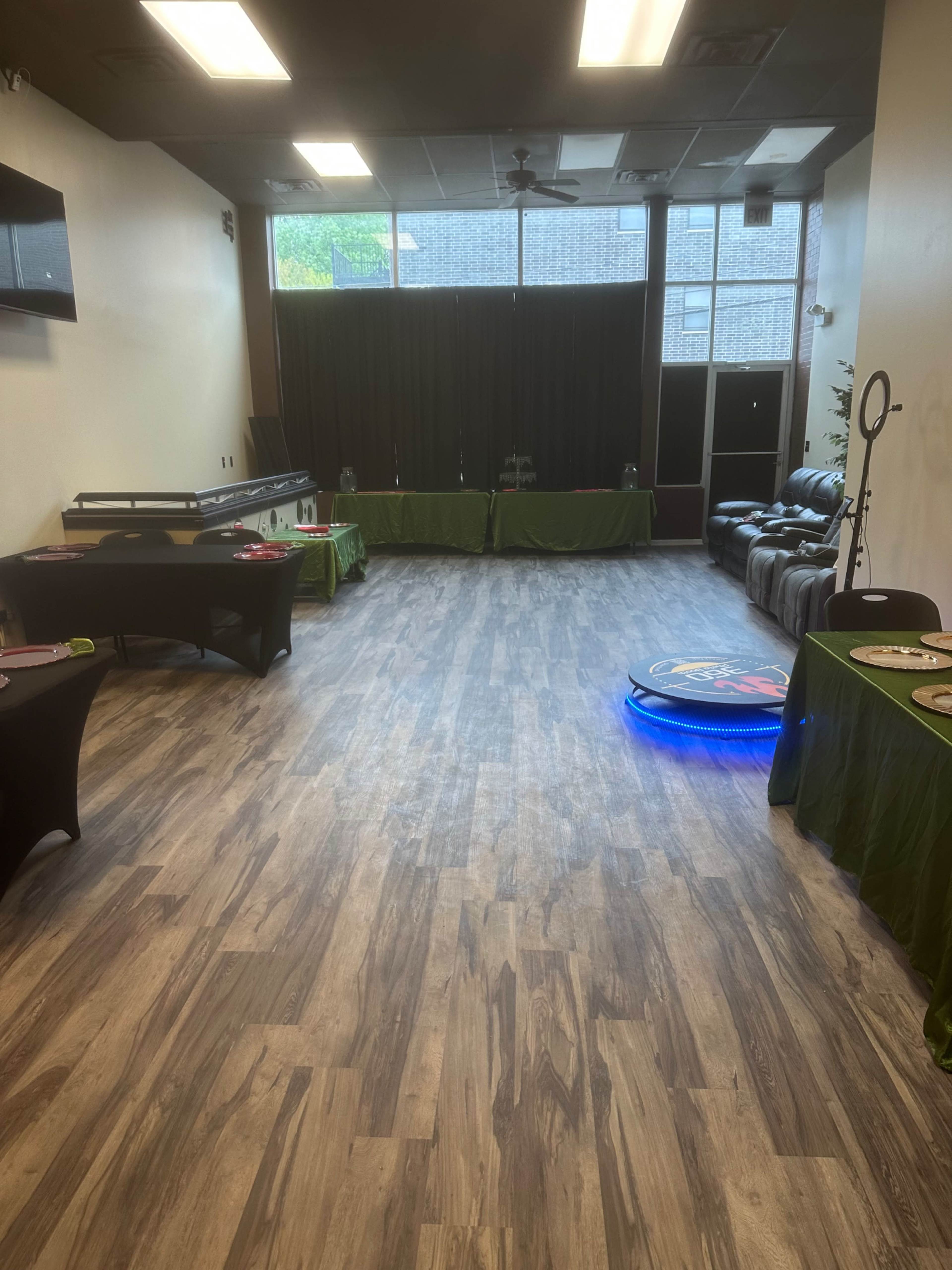 The image shows a spacious room with wooden flooring, featuring two tables covered with green cloths, a black sofa, and a colorful circular light on the floor.