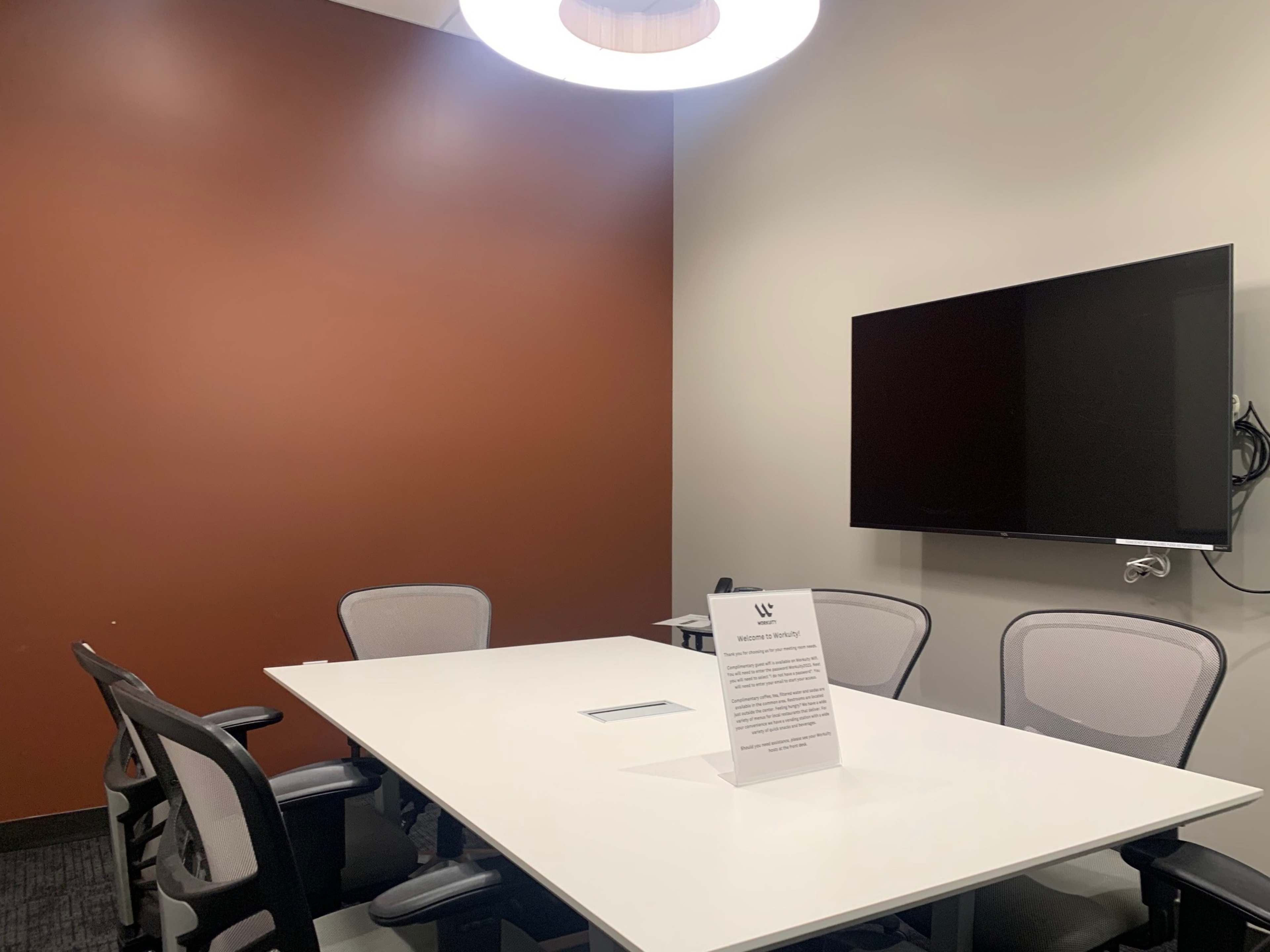 A small meeting room features a rectangular table with four chairs, a wall-mounted TV, and a sign on the table.