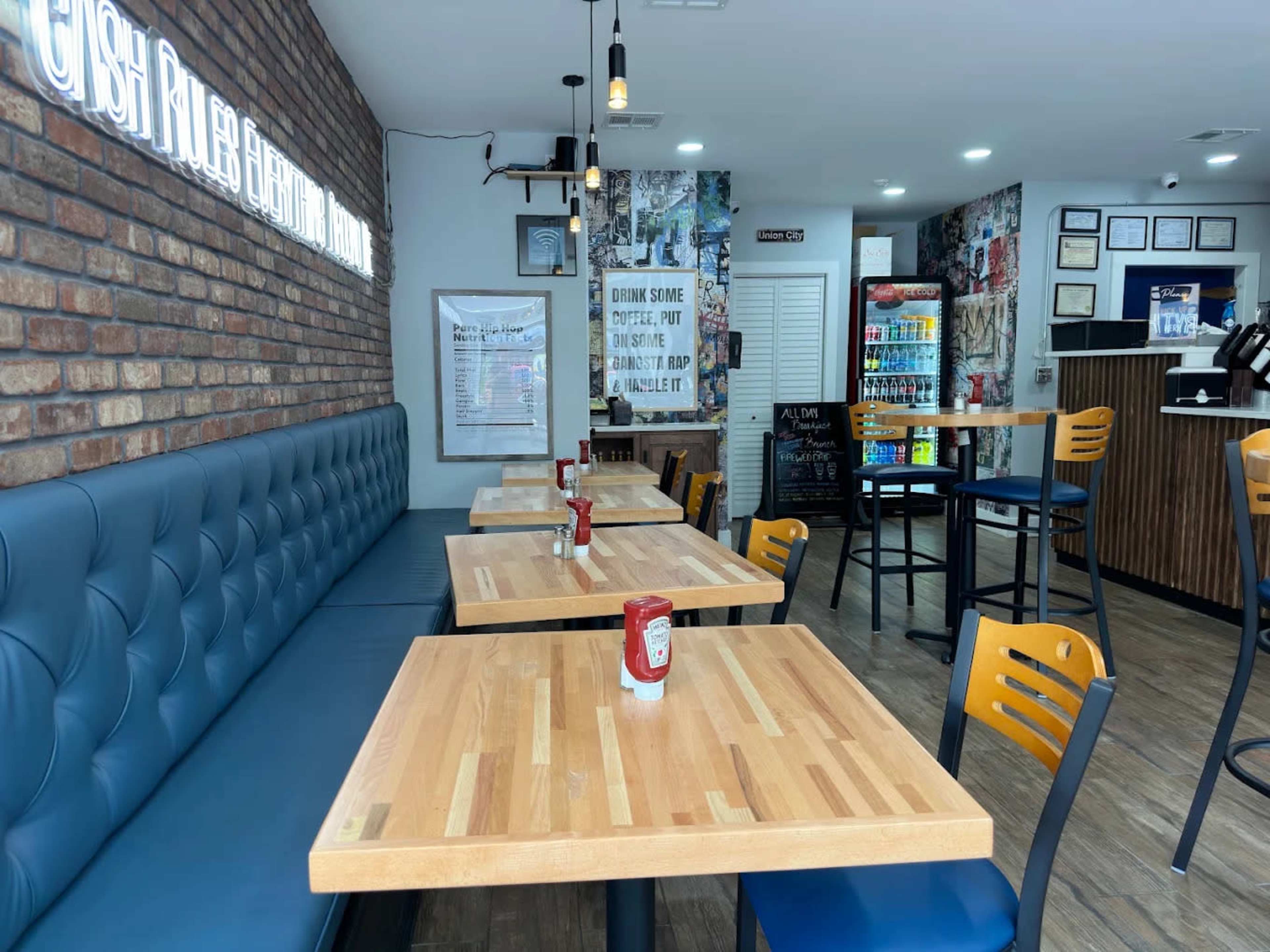 The image shows a casual dining area with wooden tables, blue upholstered seating, and a brick wall decorated with various posters and memorabilia.