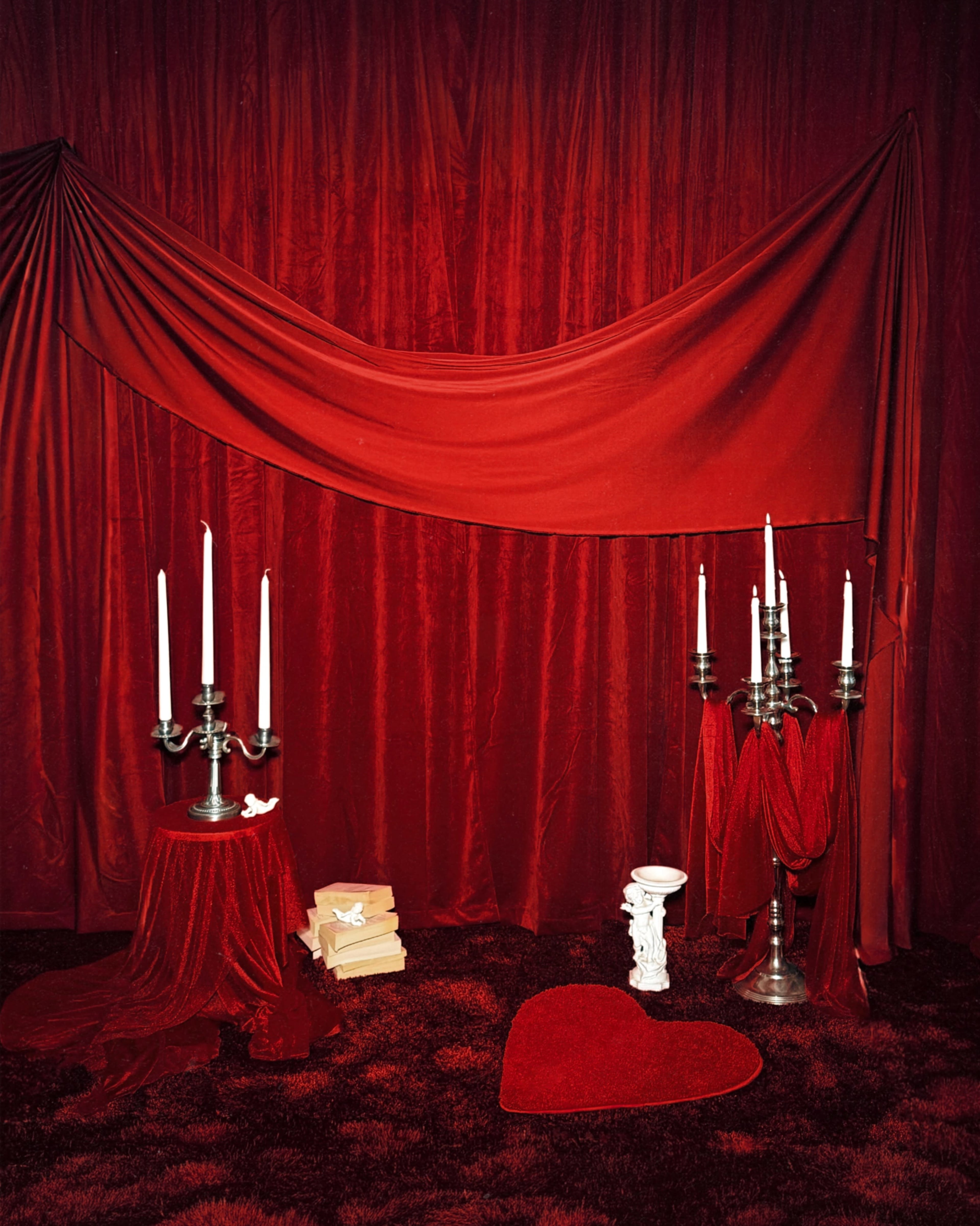 The scene features a richly draped red background with a table, candelabra, stacks of books, and a heart-shaped rug on a plush floor.