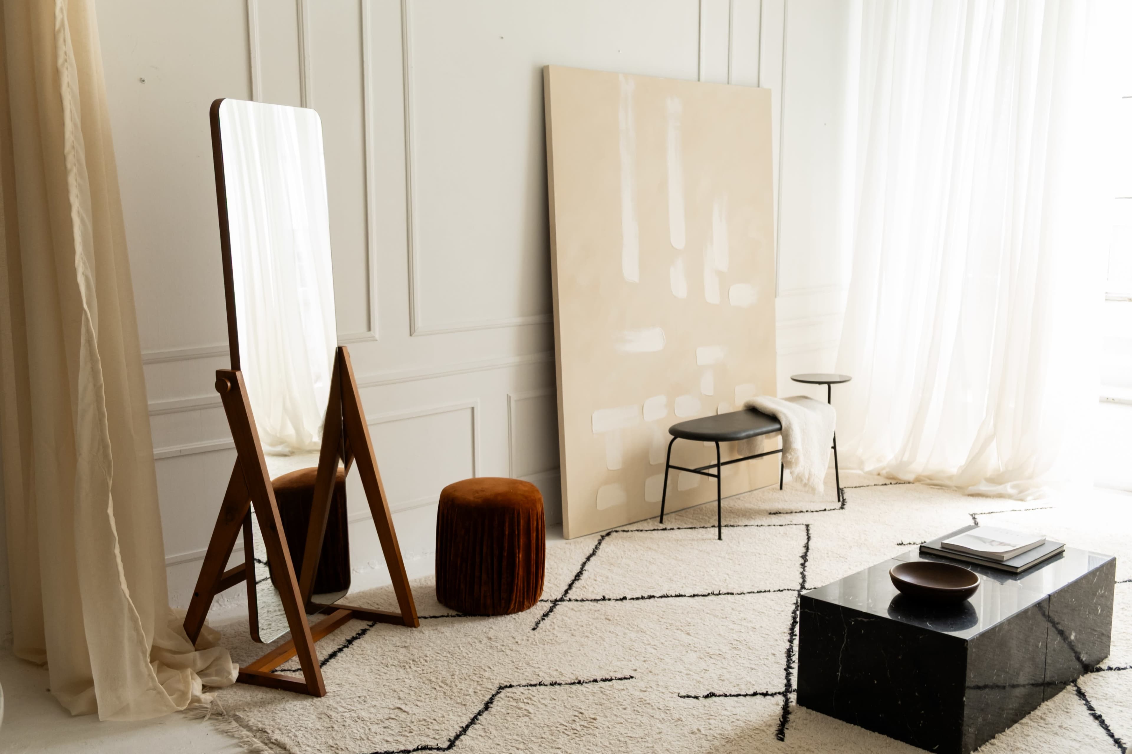 The scene features a minimalist interior with a wooden mirror, an orange pouf, a black coffee table, and a large piece of art against a light-colored wall.