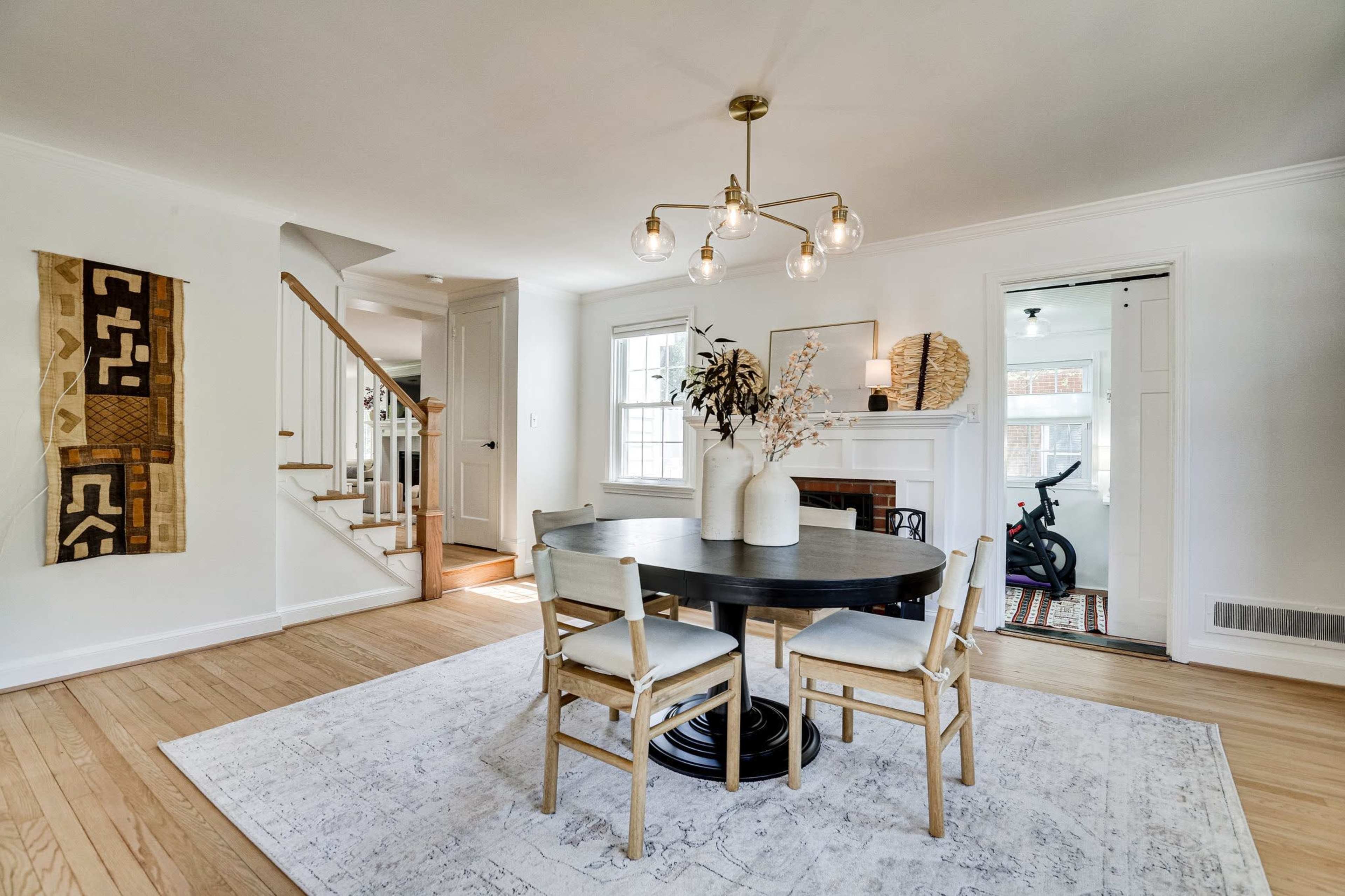 A round dining table with four chairs is set in a well-lit room featuring a staircase, a modern light fixture, and decorative elements on the walls.