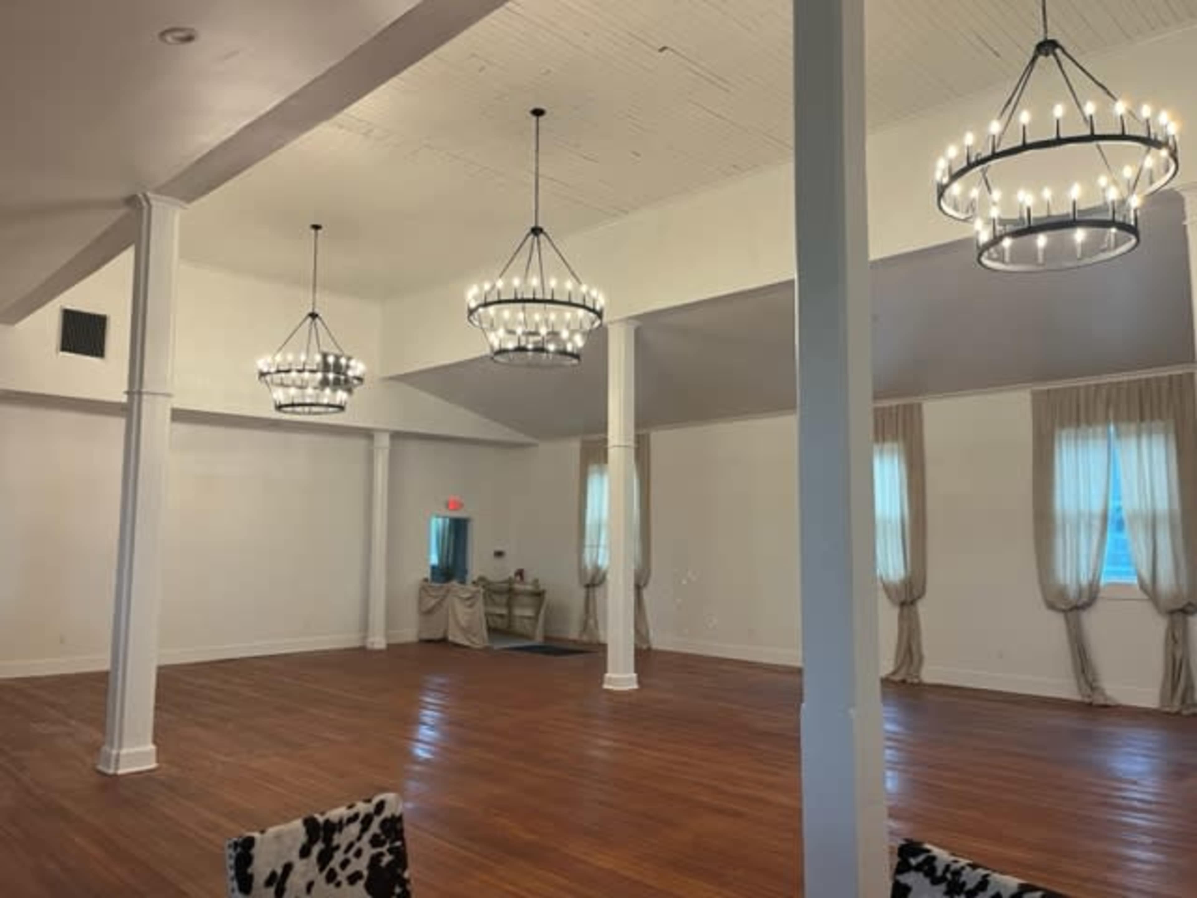 A spacious indoor venue with wooden floors, tall white walls, and three large chandeliers hanging from the ceiling.