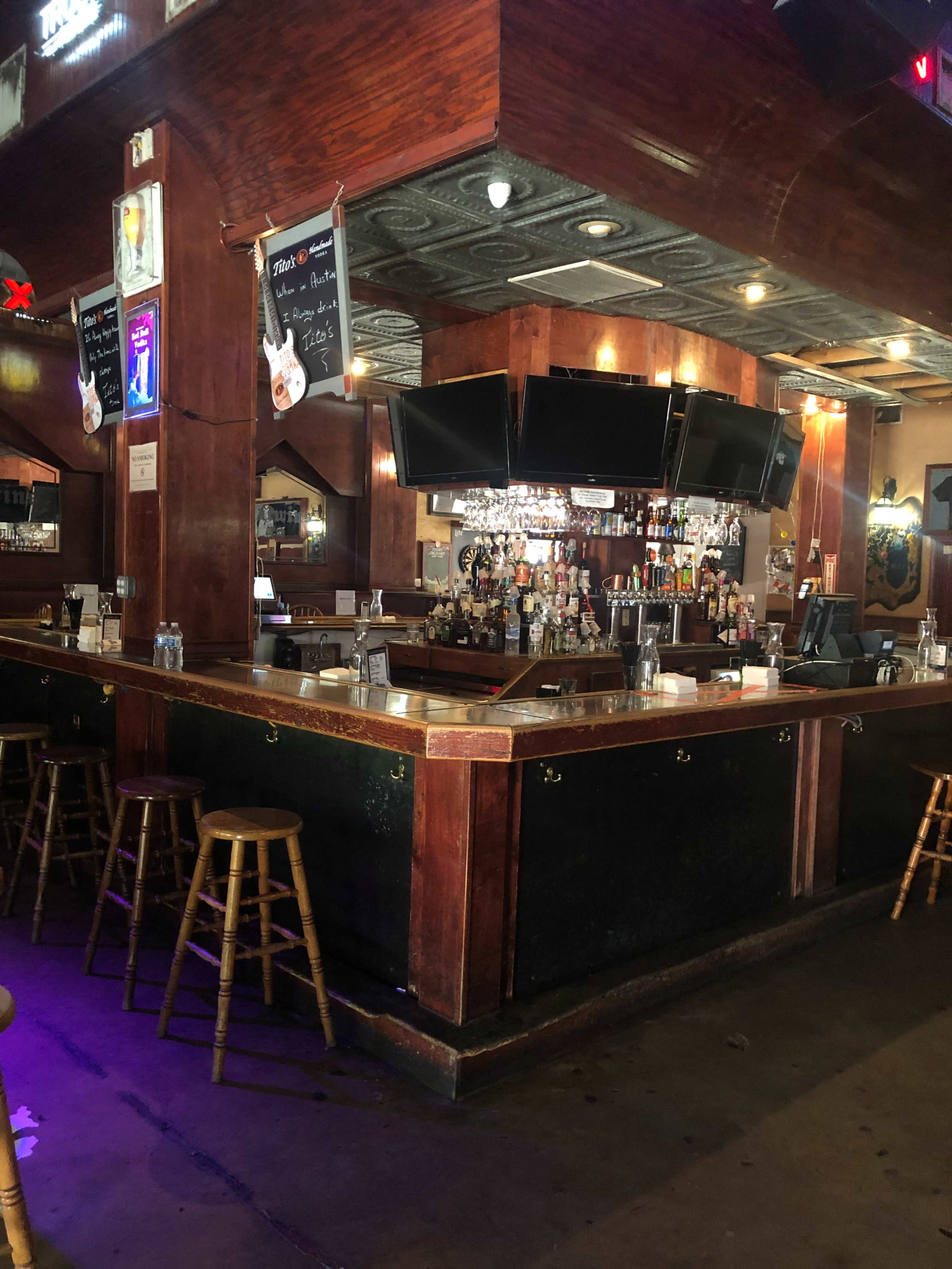 The image shows a bar area with wooden counters, high stools, and multiple screens mounted above the bar, displaying a variety of bottles and a chandelier in the background.
