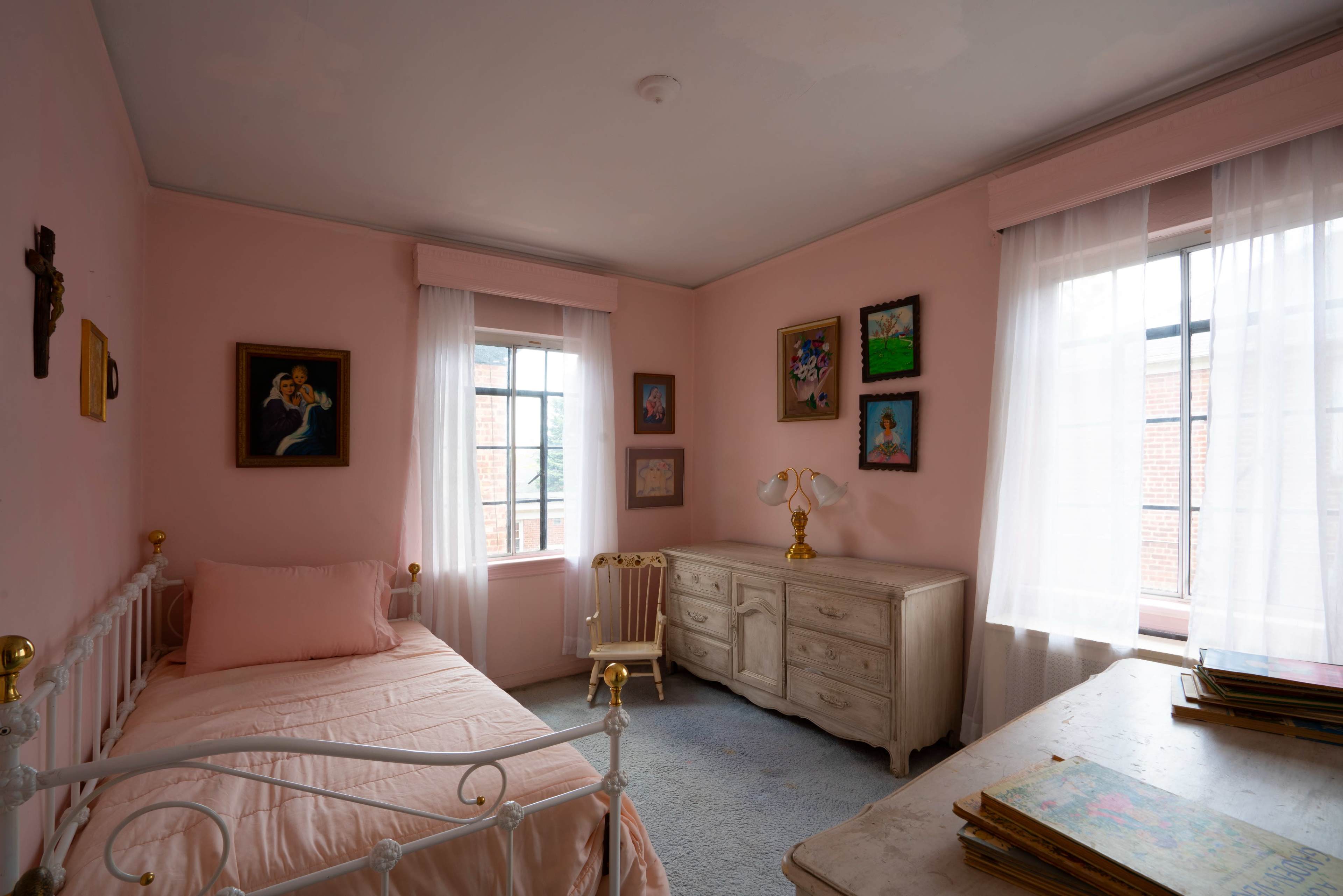 The room features pink walls, a bed with a light pink cover, a wooden dresser, and several framed paintings, all illuminated by natural light through the windows.