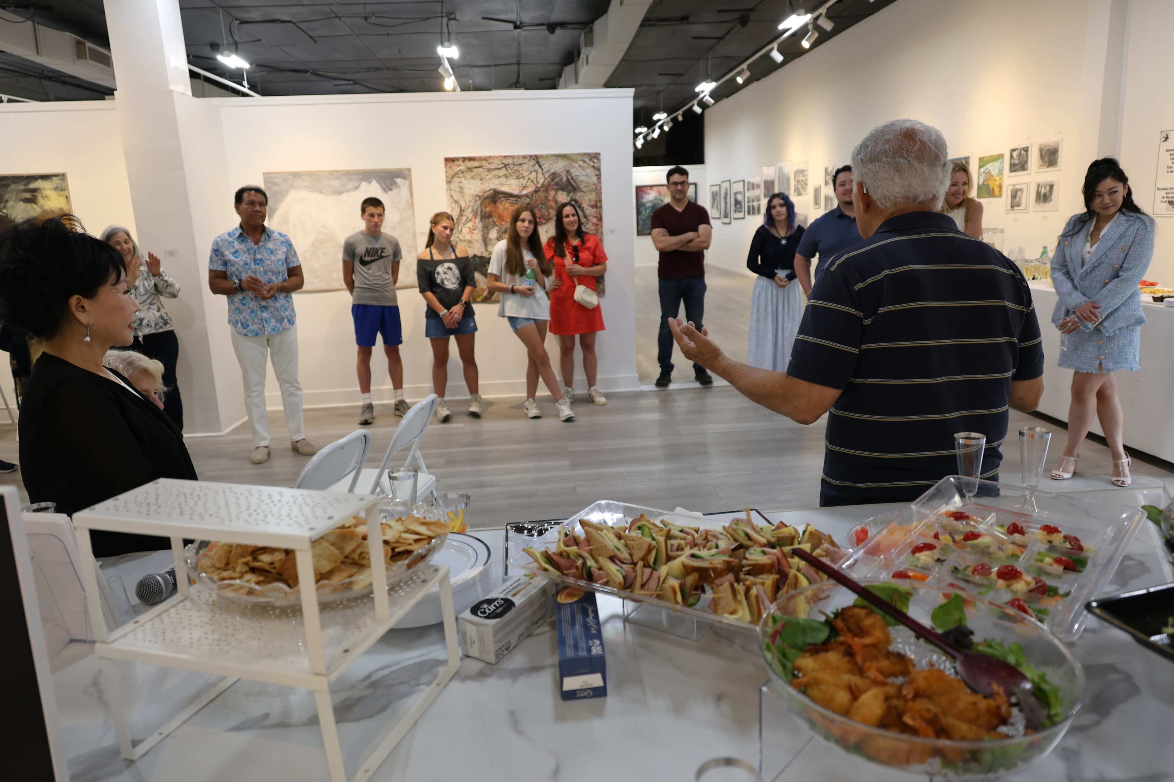 A group of people watches a speaker in an art gallery, where a variety of hors d'oeuvres are displayed on a table.