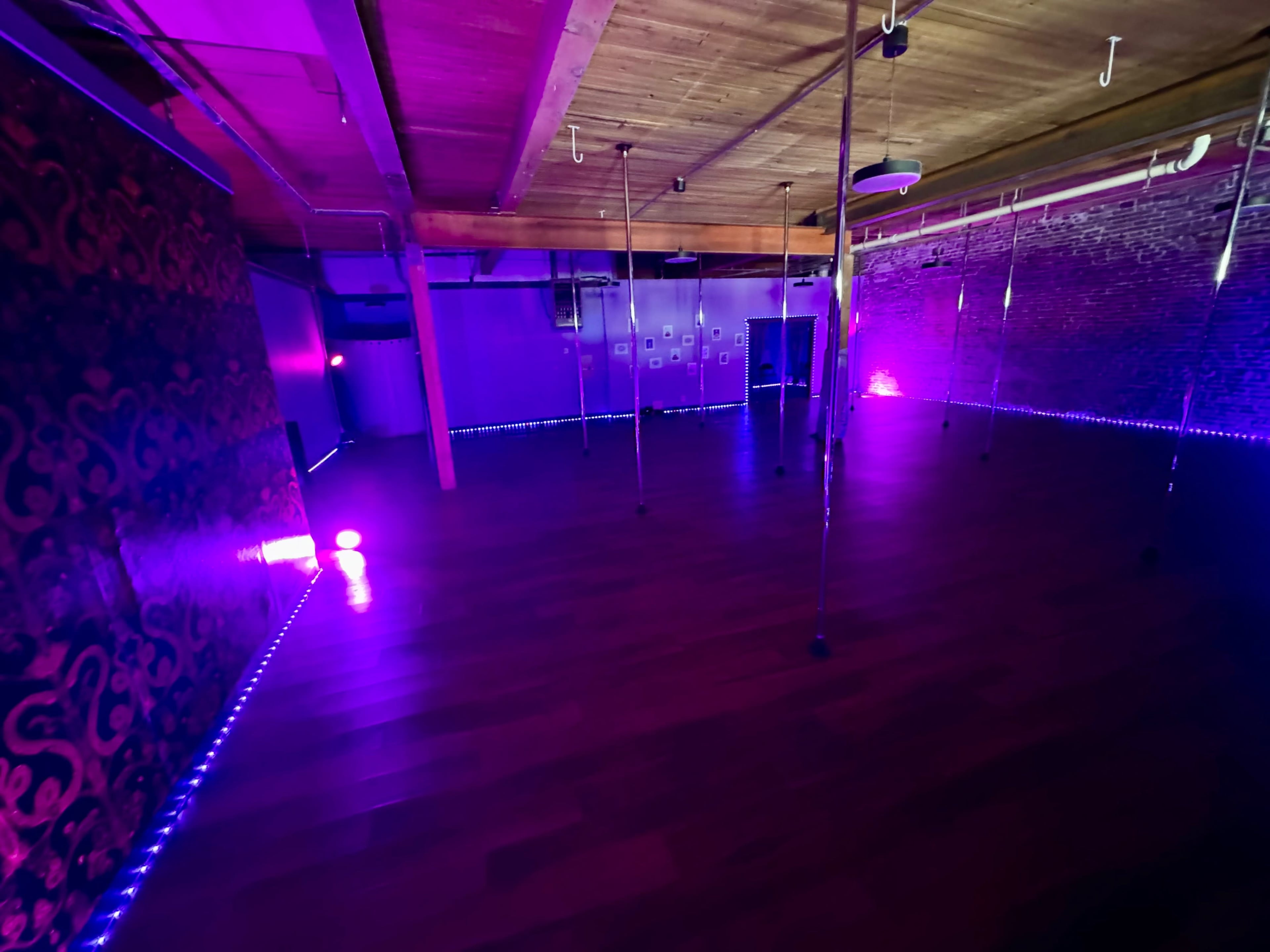 The image shows a dimly lit dance studio with wooden floors and several vertical poles, illuminated by purple LED lights along the walls.