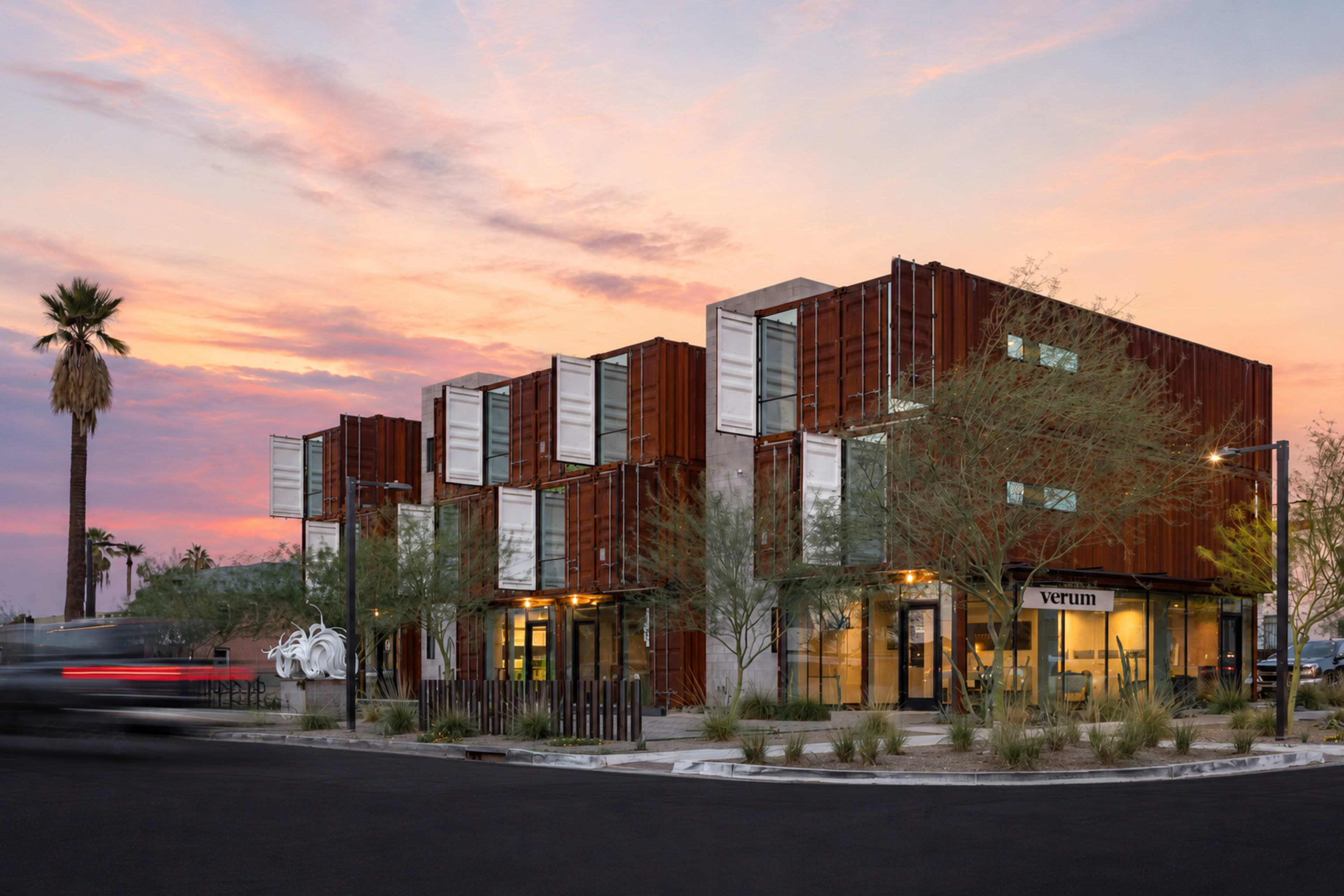 This image shows a modern commercial building constructed from shipping containers, surrounded by desert landscaping and palm trees, set against a colorful sunset sky.