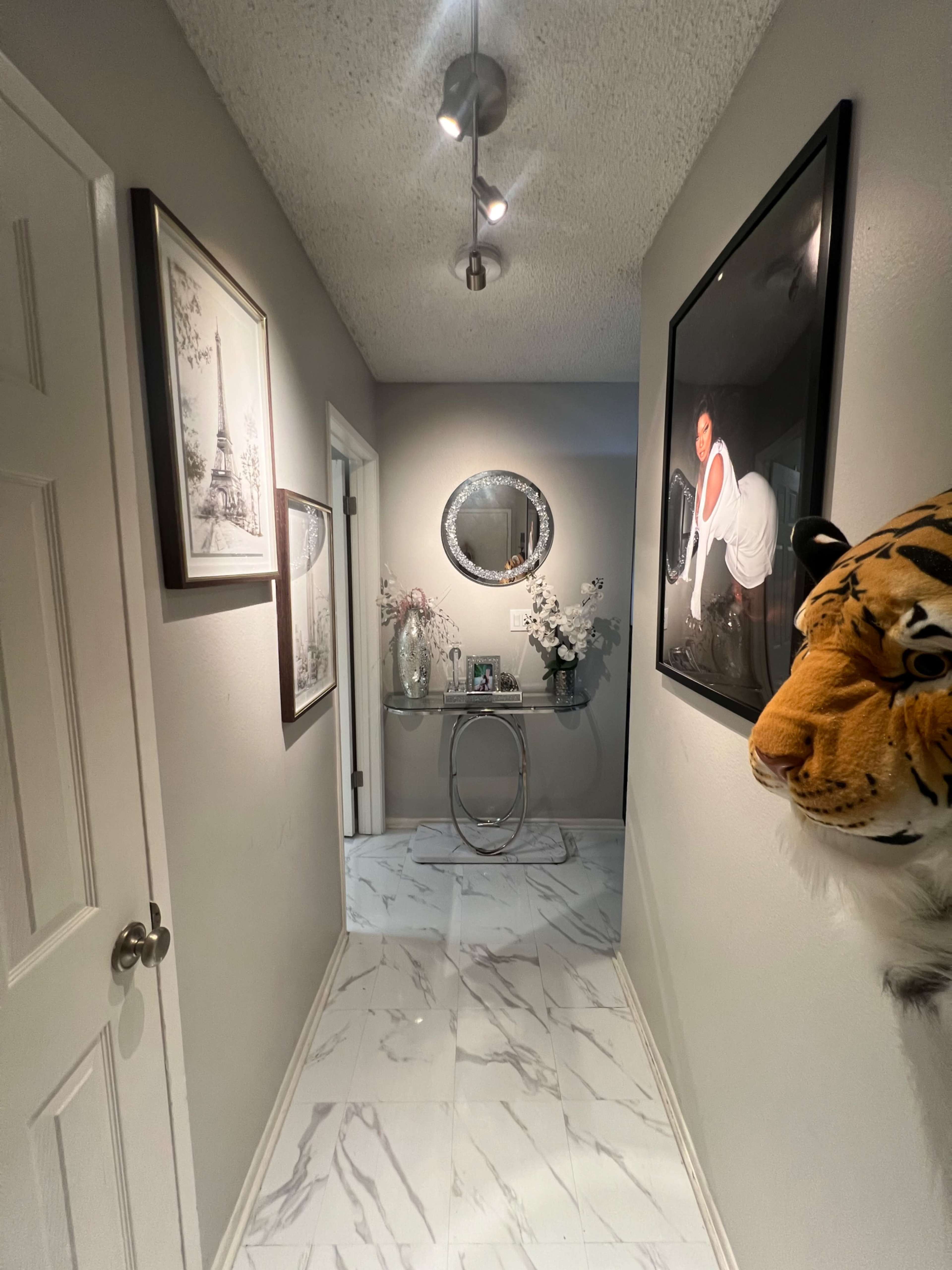 A hallway featuring marble-patterned flooring, decorative wall art, and a round mirror above a console table.