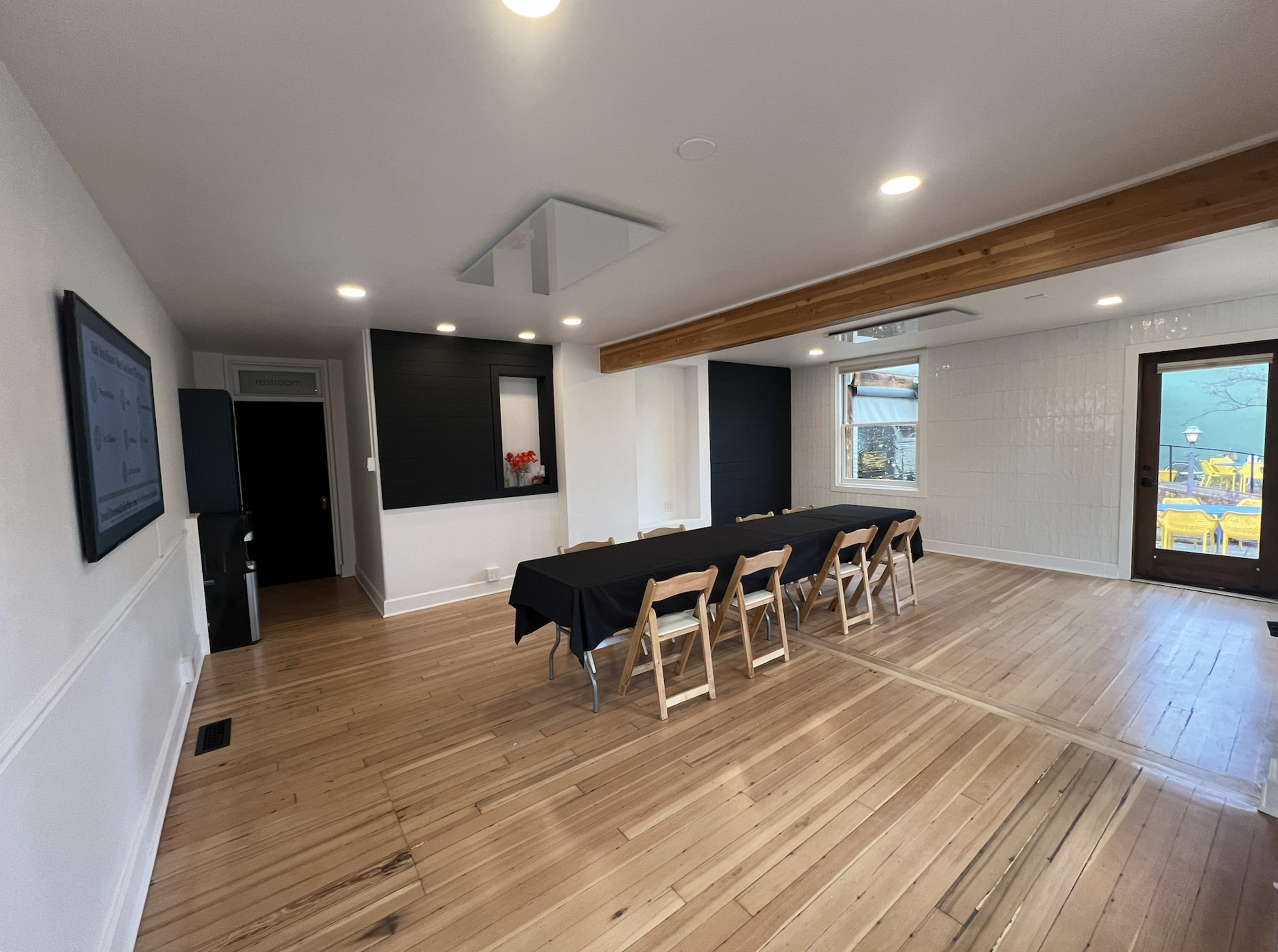 The image depicts a modern meeting room with a long black table surrounded by wooden chairs, hardwood floors, and large windows allowing natural light.