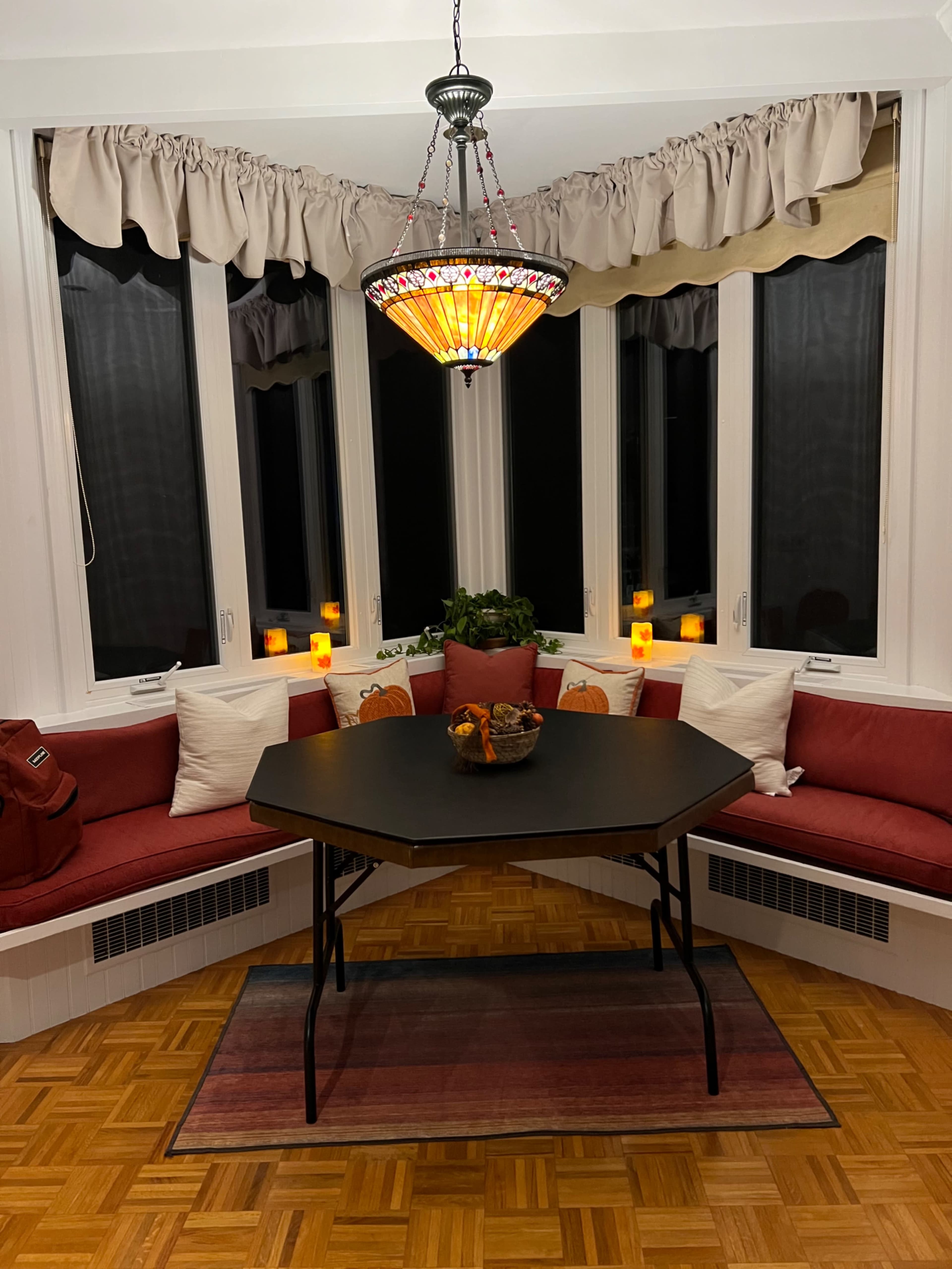 A round table with a black surface is set in a bay window surrounded by cushioned seating and illuminated by a stained glass chandelier.