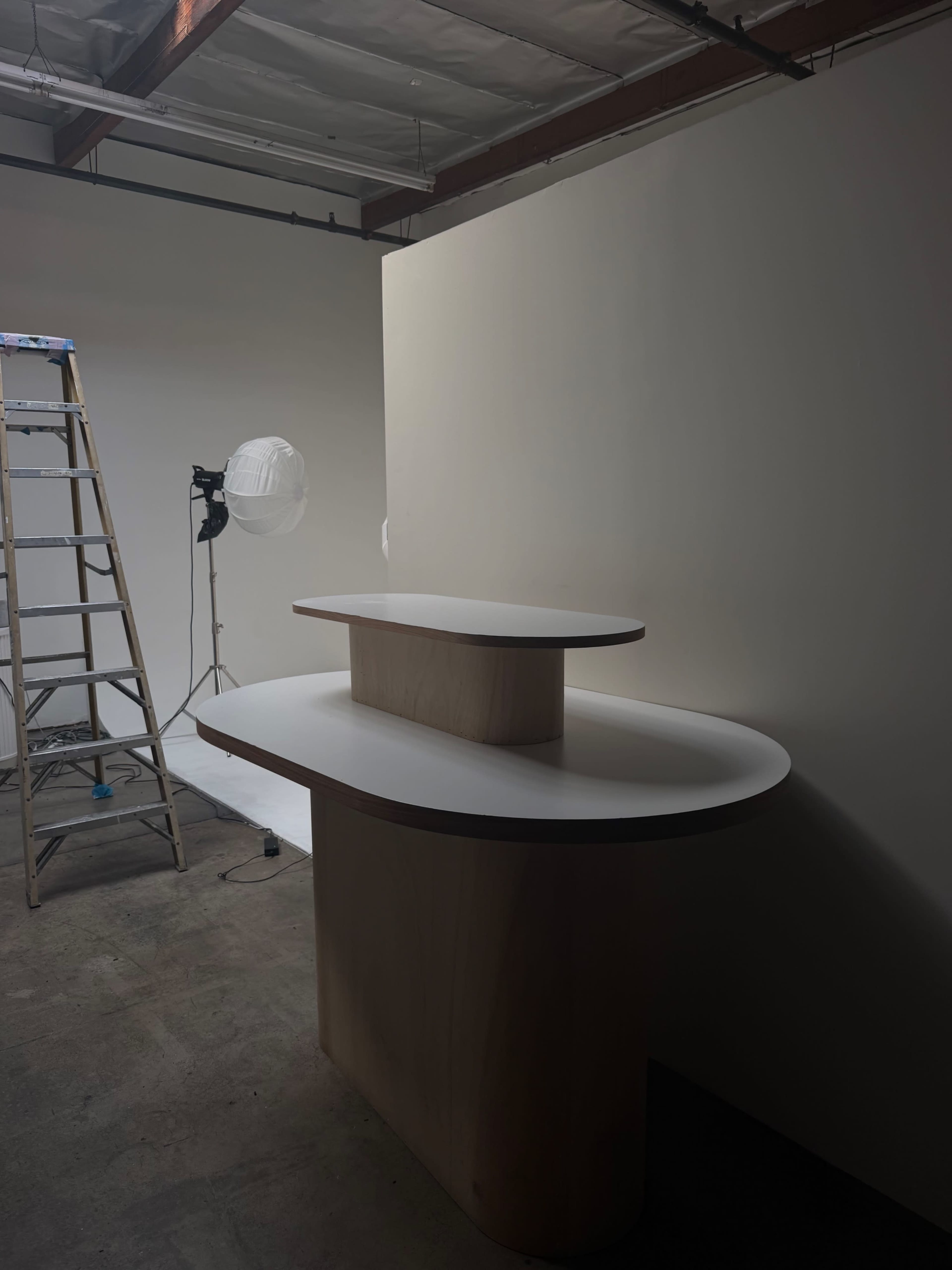 The image shows a minimalistic countertop setup with a light-colored wooden finish, accompanied by a ladder and a light stand in an empty room.