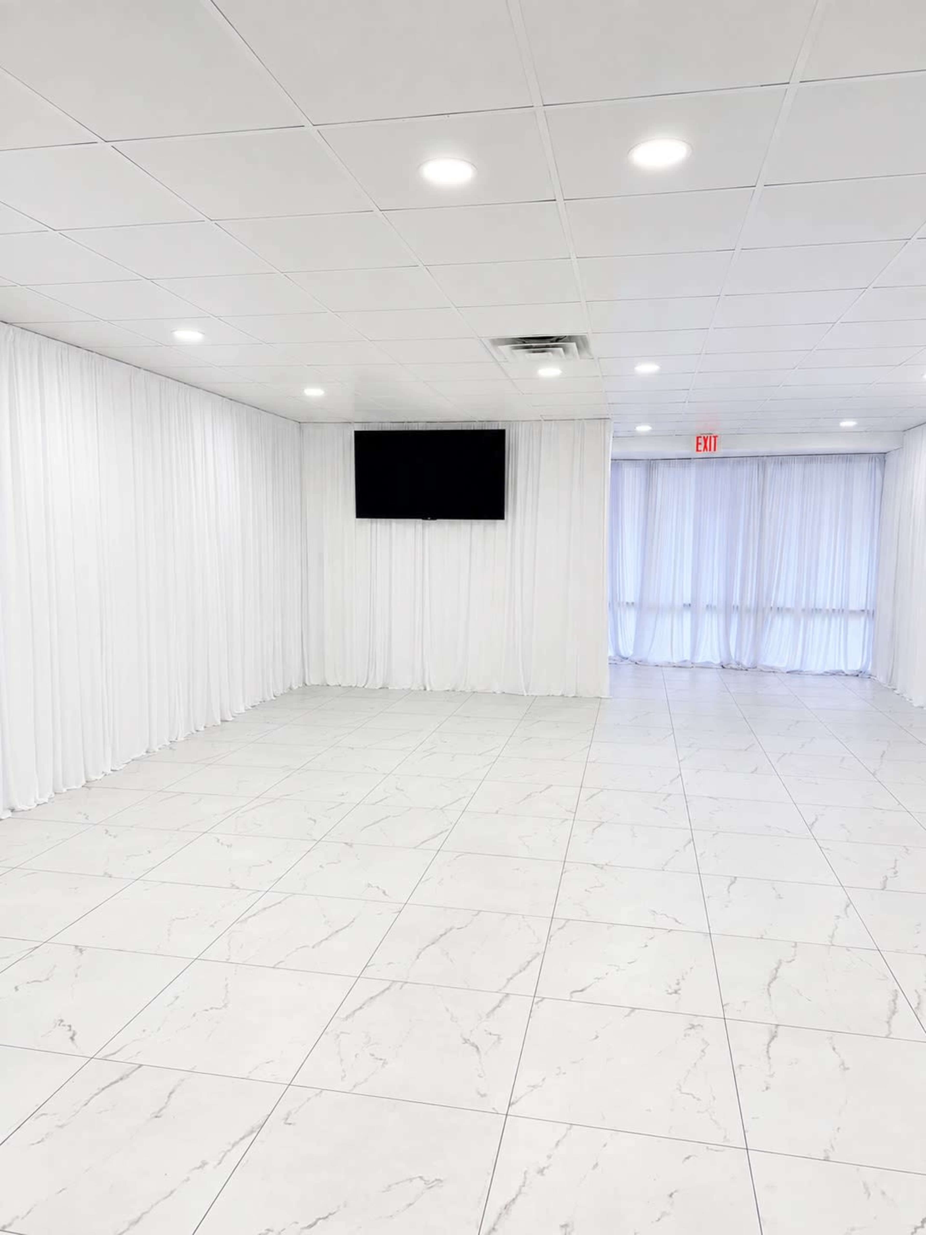 The image shows a spacious, well-lit room with white walls and draped curtains, featuring a television mounted on one wall and large windows covered by sheer curtains.