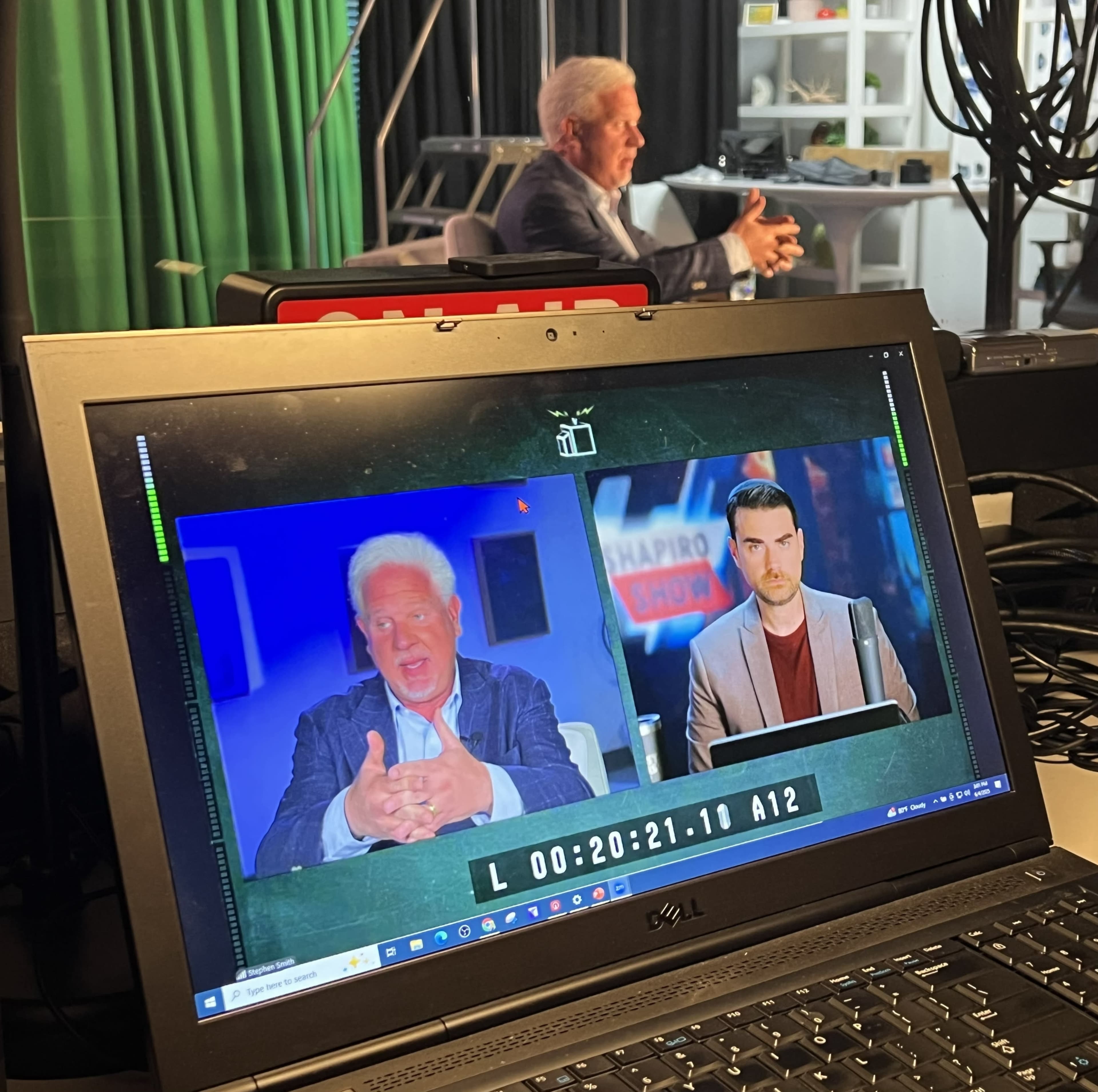 A computer monitor displays a split screen with two men discussing, while a third man gestures in the background against a green screen set.