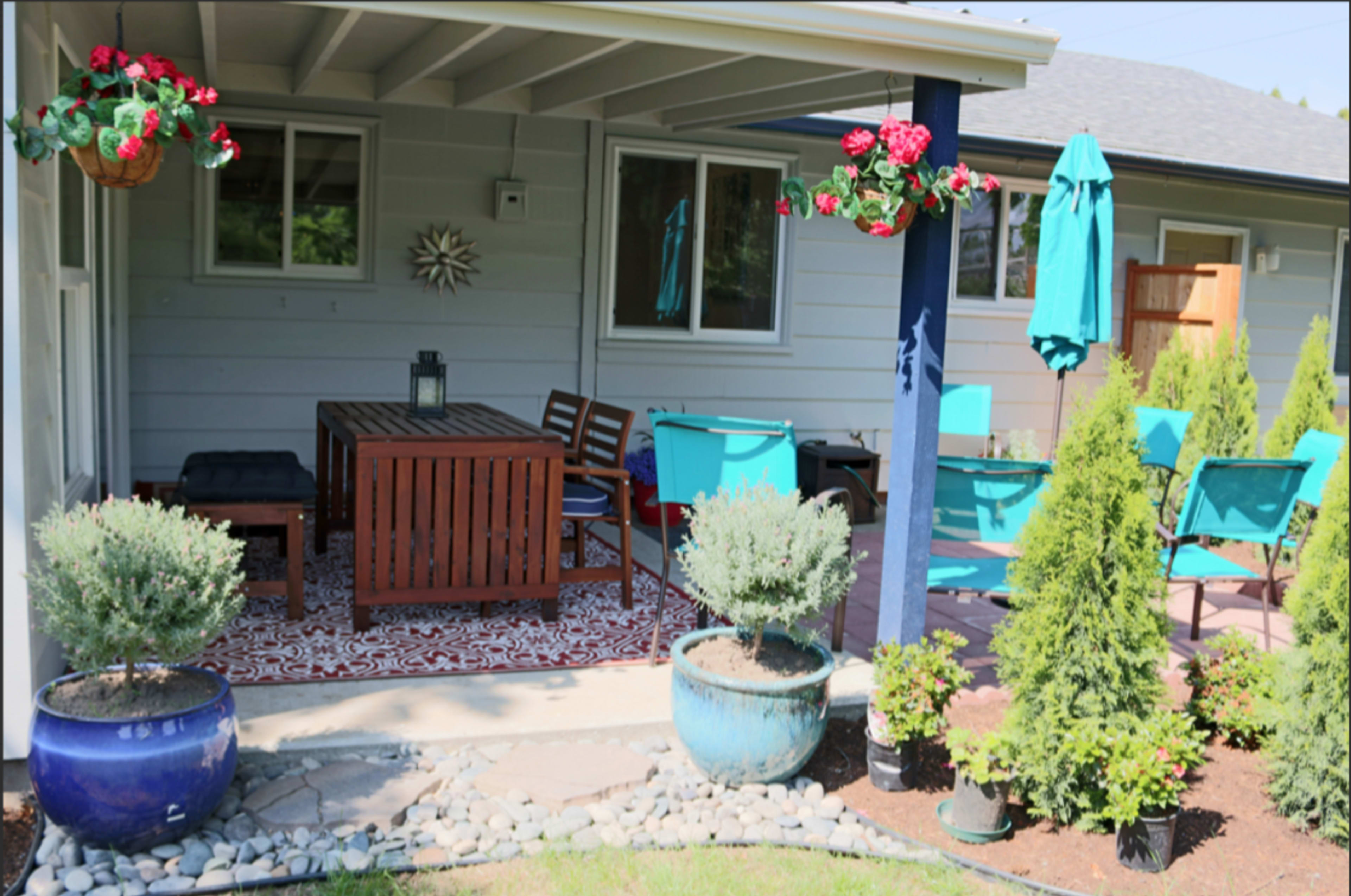 A covered patio area features a wooden dining table with chairs, potted plants, and decorative hanging flowers.