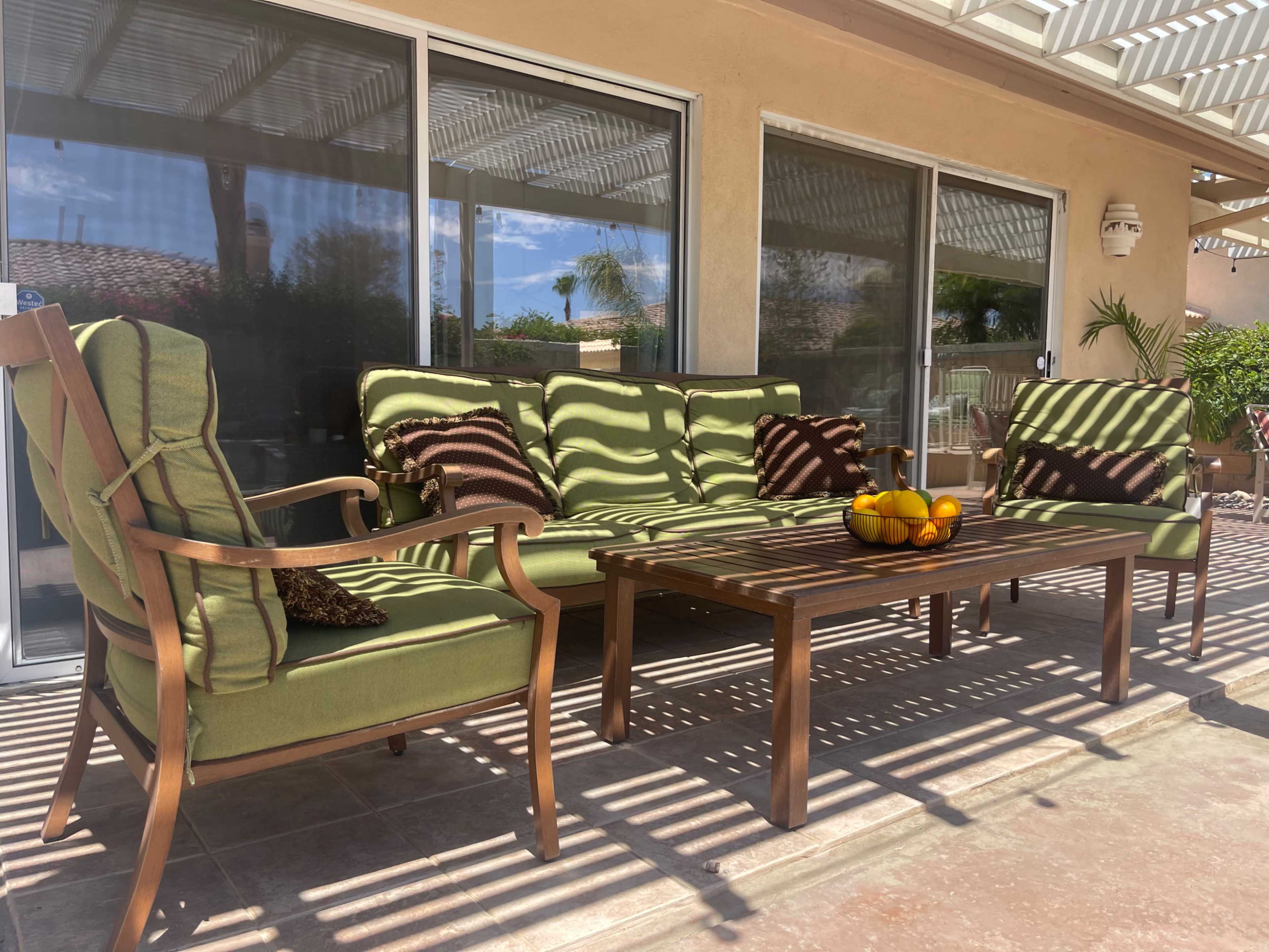 A patio seating area features green cushioned furniture arranged around a wooden table adorned with a bowl of fruit, all under a shaded pergola.