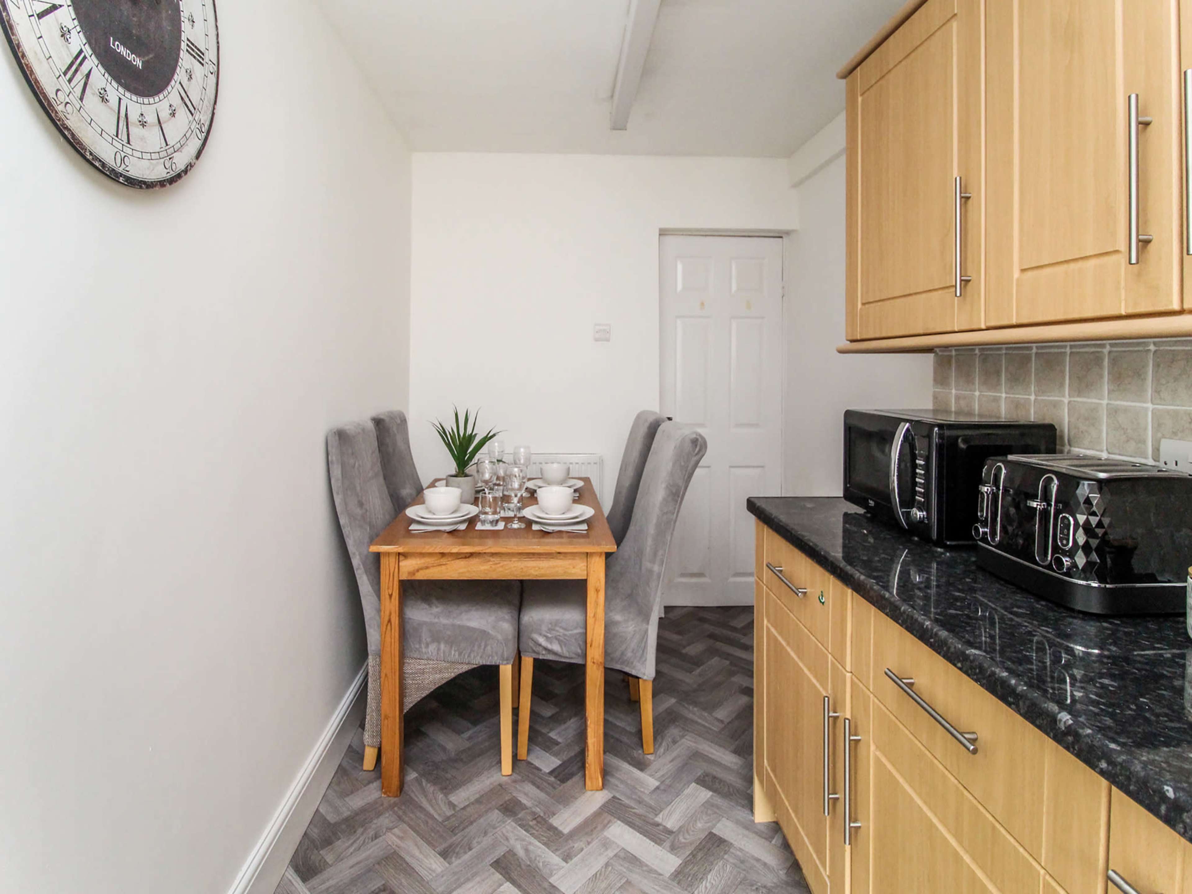 A small kitchen features a wooden table with gray upholstered chairs, set for a meal, along with a black countertop and kitchen appliances.