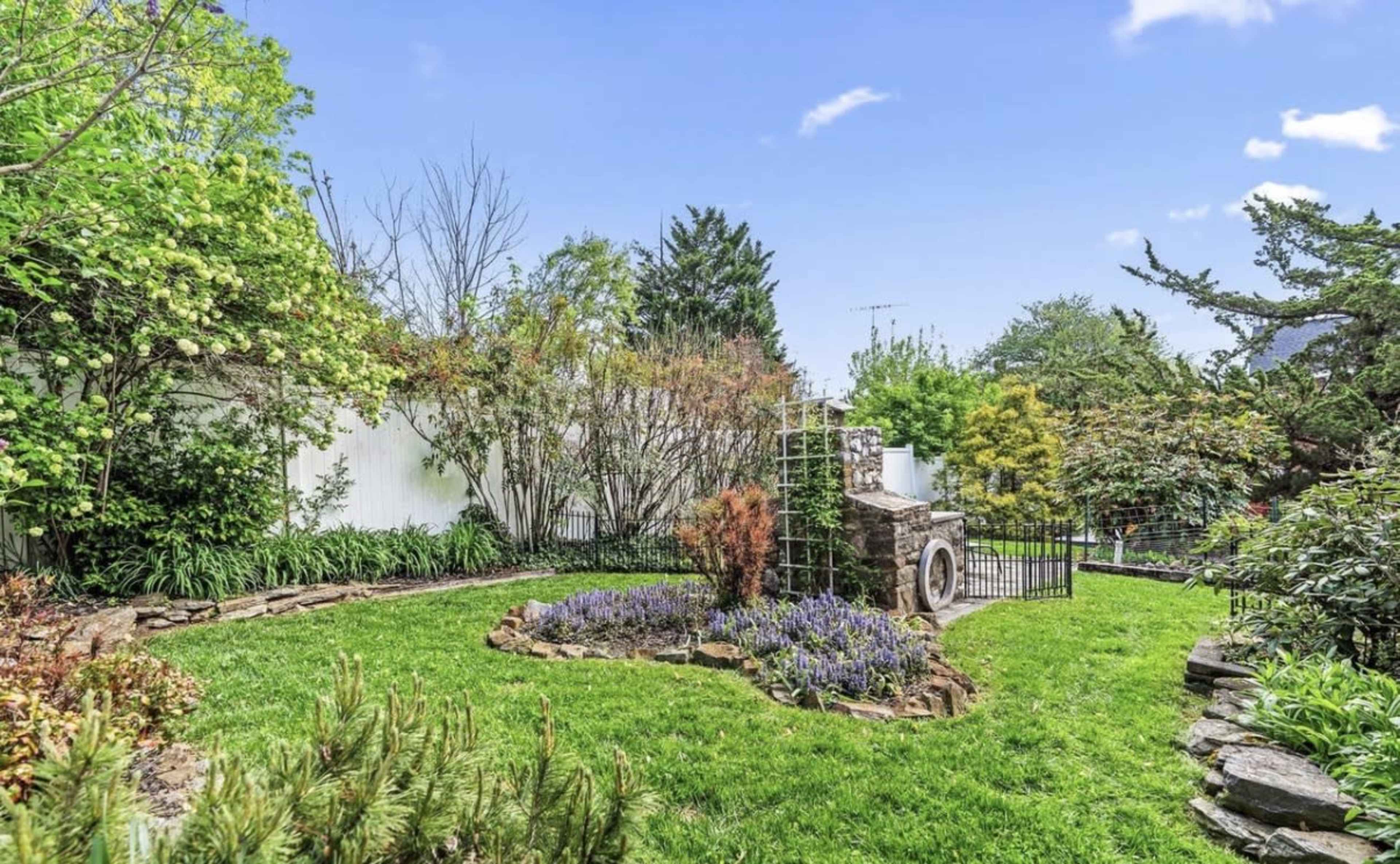 The image shows a well-maintained garden featuring a mix of greenery, blooming flowers, and a stone feature with a gate.