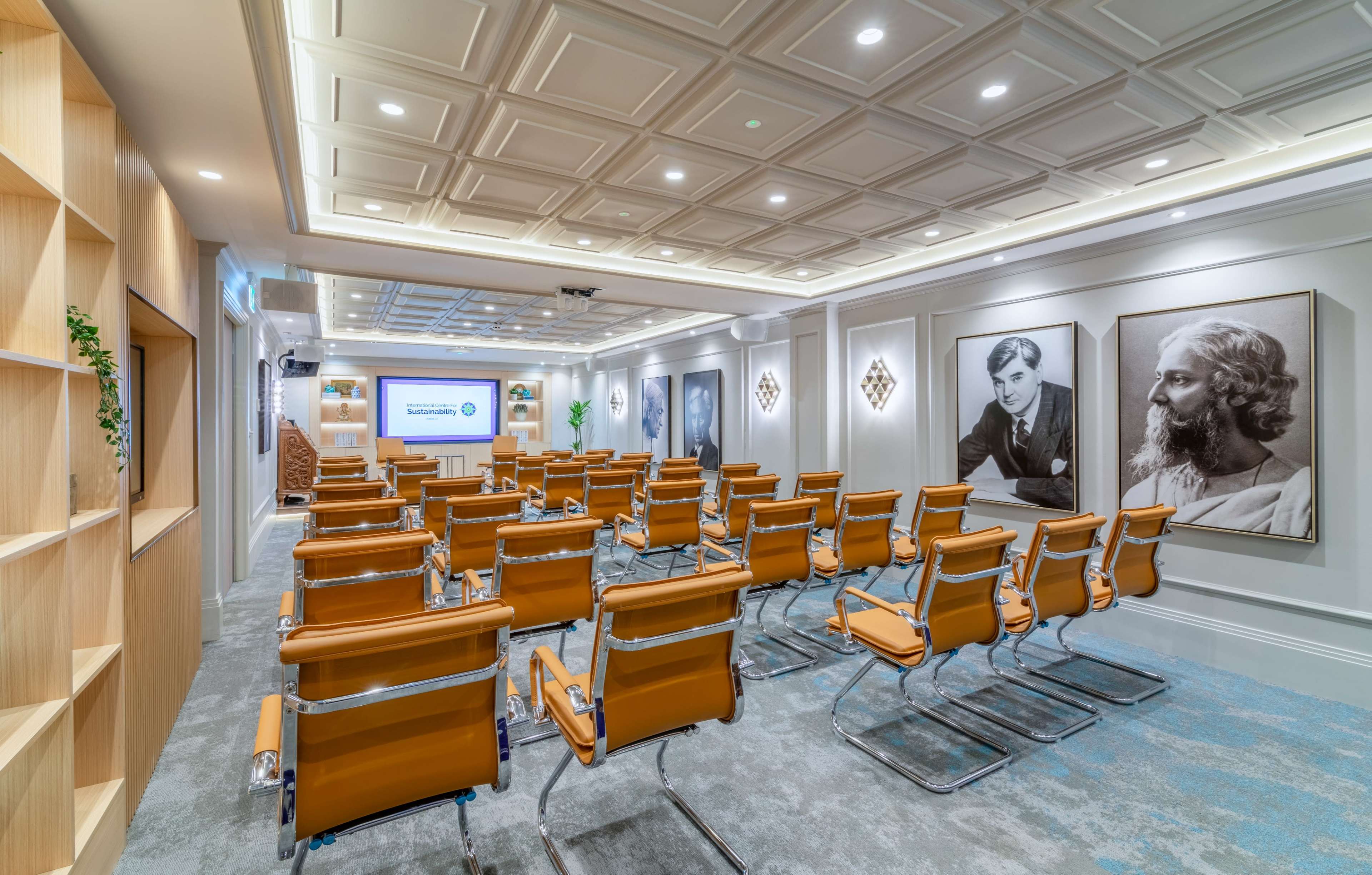 The image shows a conference room with orange and silver chairs arranged in rows, featuring two large framed portraits on the walls and a presentation screen at the front.