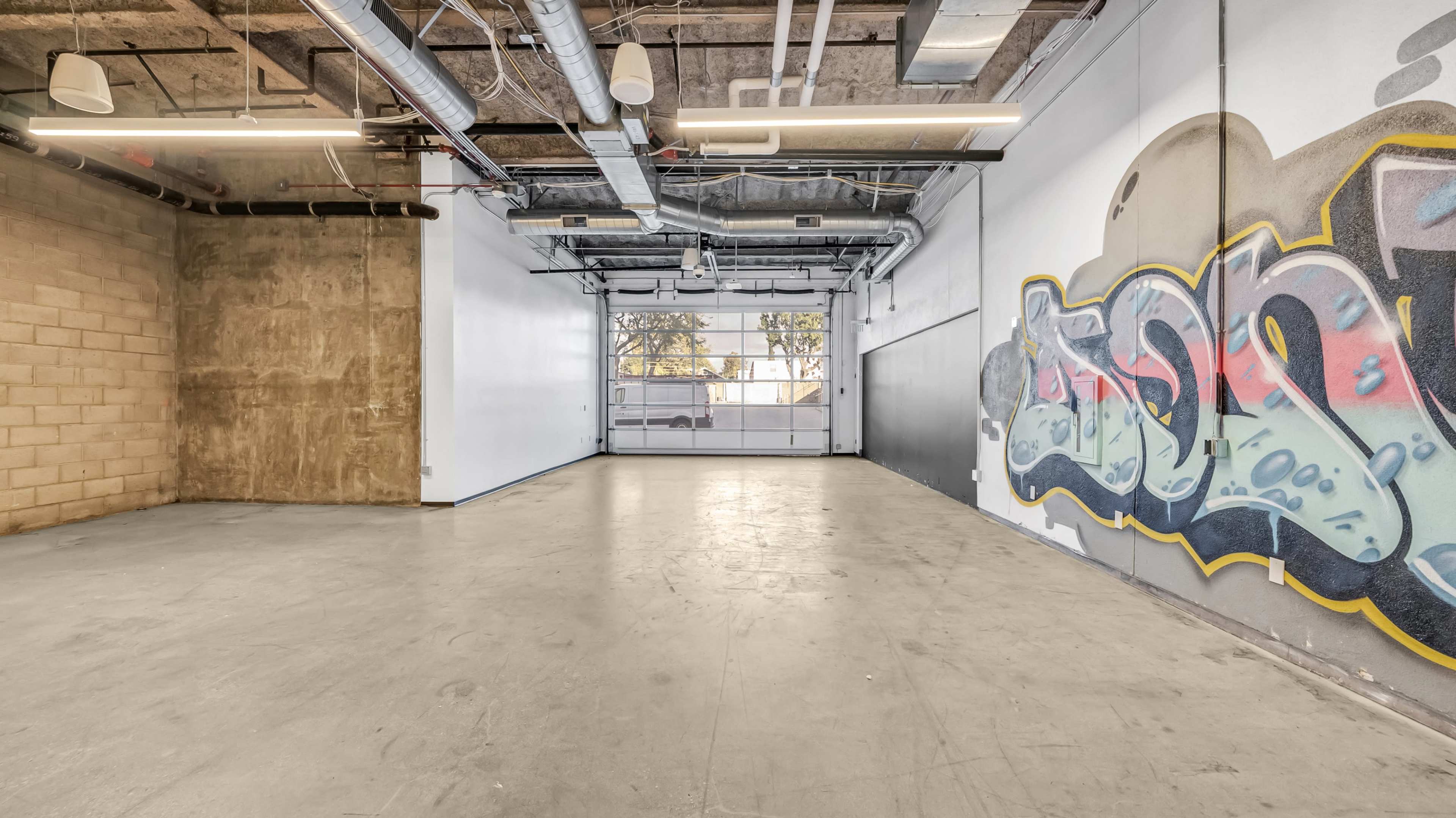 An empty industrial space featuring a concrete floor and a large wall with graffiti art.
