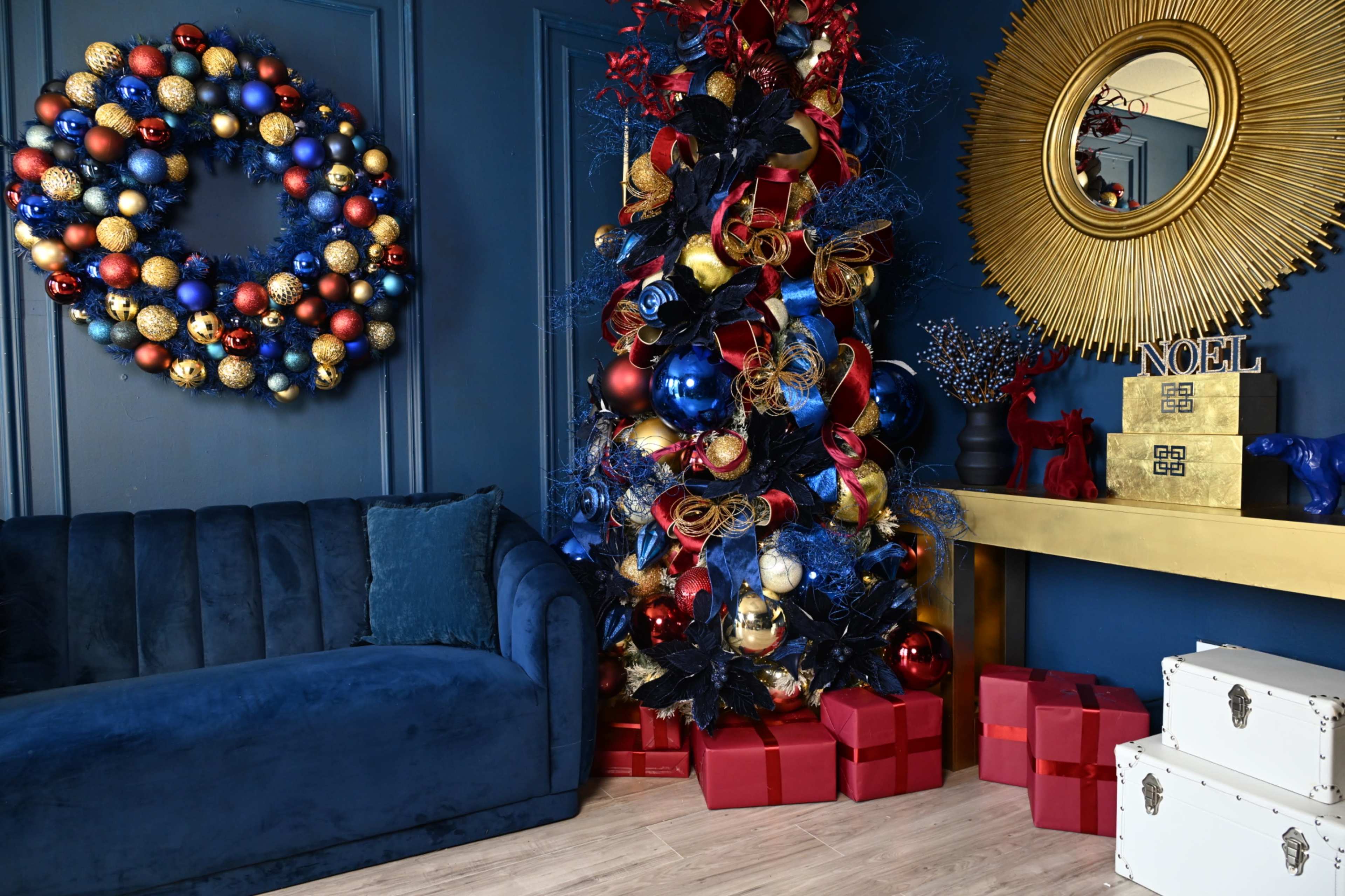 The scene features a decorated living room with a blue couch, a Christmas tree adorned with colorful ornaments, a golden circular mirror, and a collection of wrapped gifts.