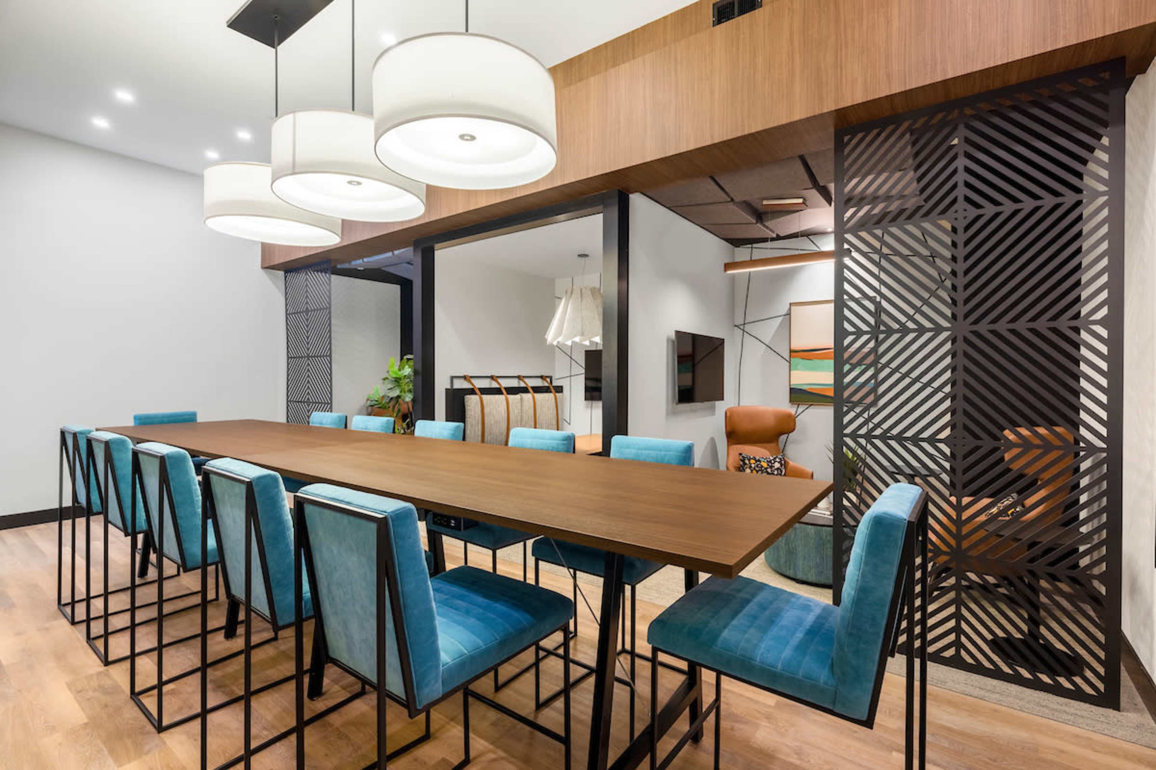 A long wooden table is surrounded by blue upholstered chairs in a modern dining area with pendant lighting and decorative wall panels.