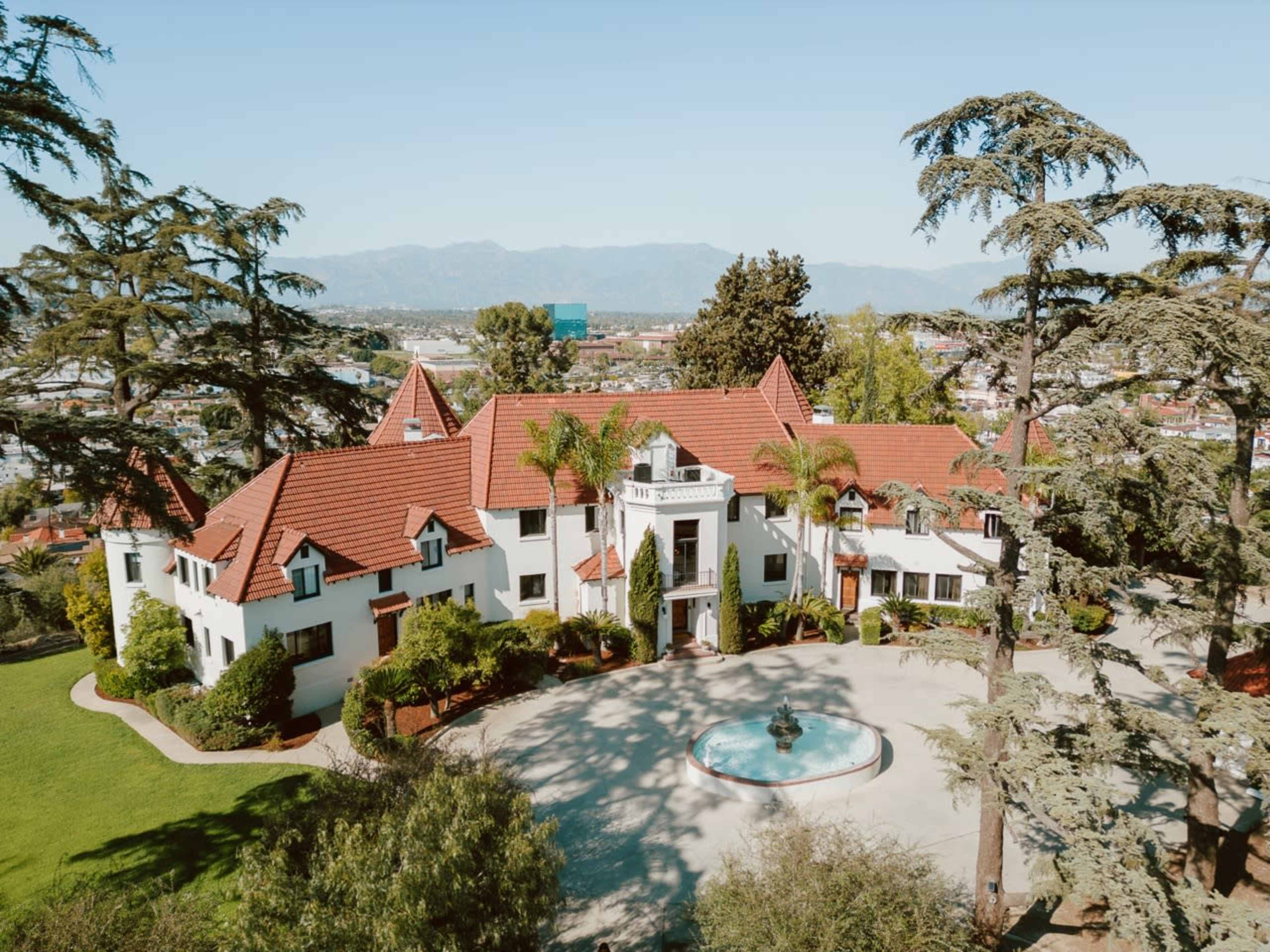 A large, white mansion with a red-tiled roof, surrounded by greenery and a circular driveway with a fountain in the front.