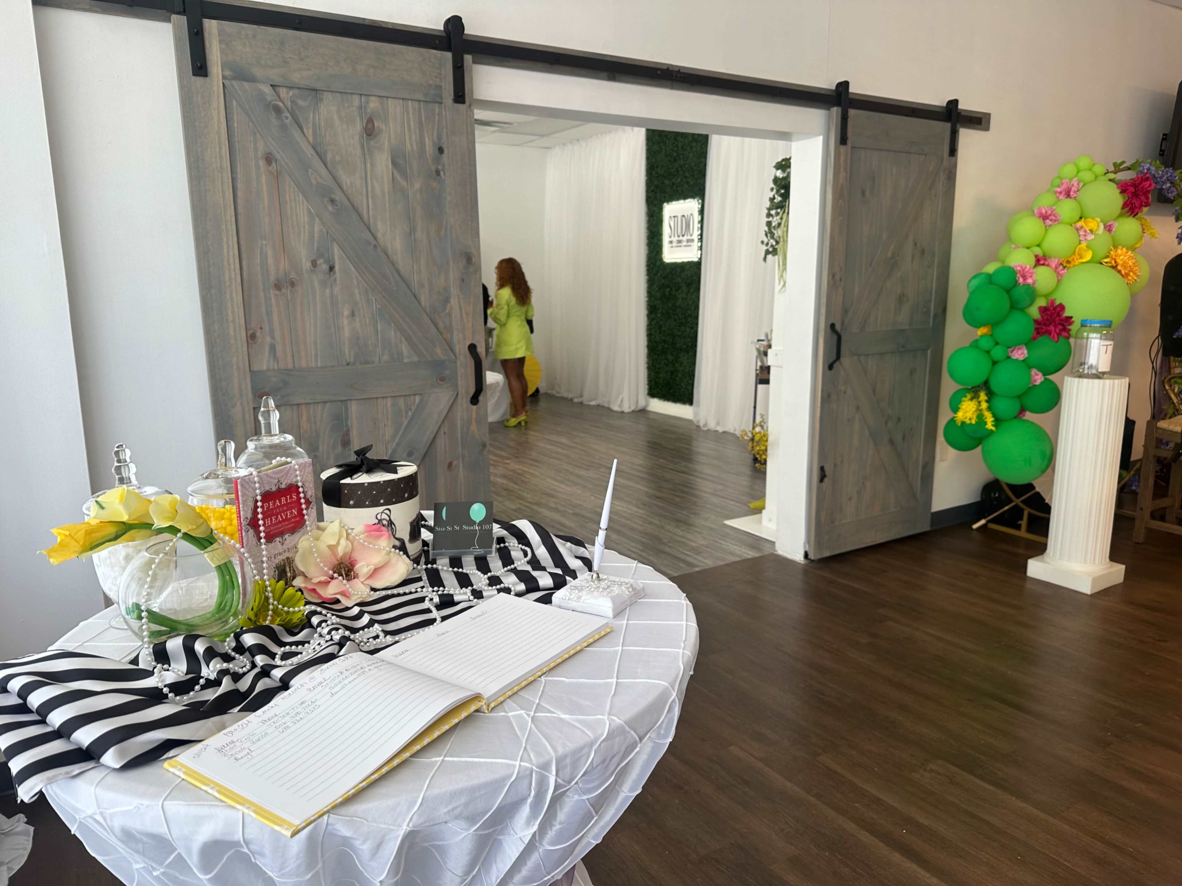A bright event space features sliding barn doors, a decorated table with a guest book, and a backdrop adorned with colorful balloons.