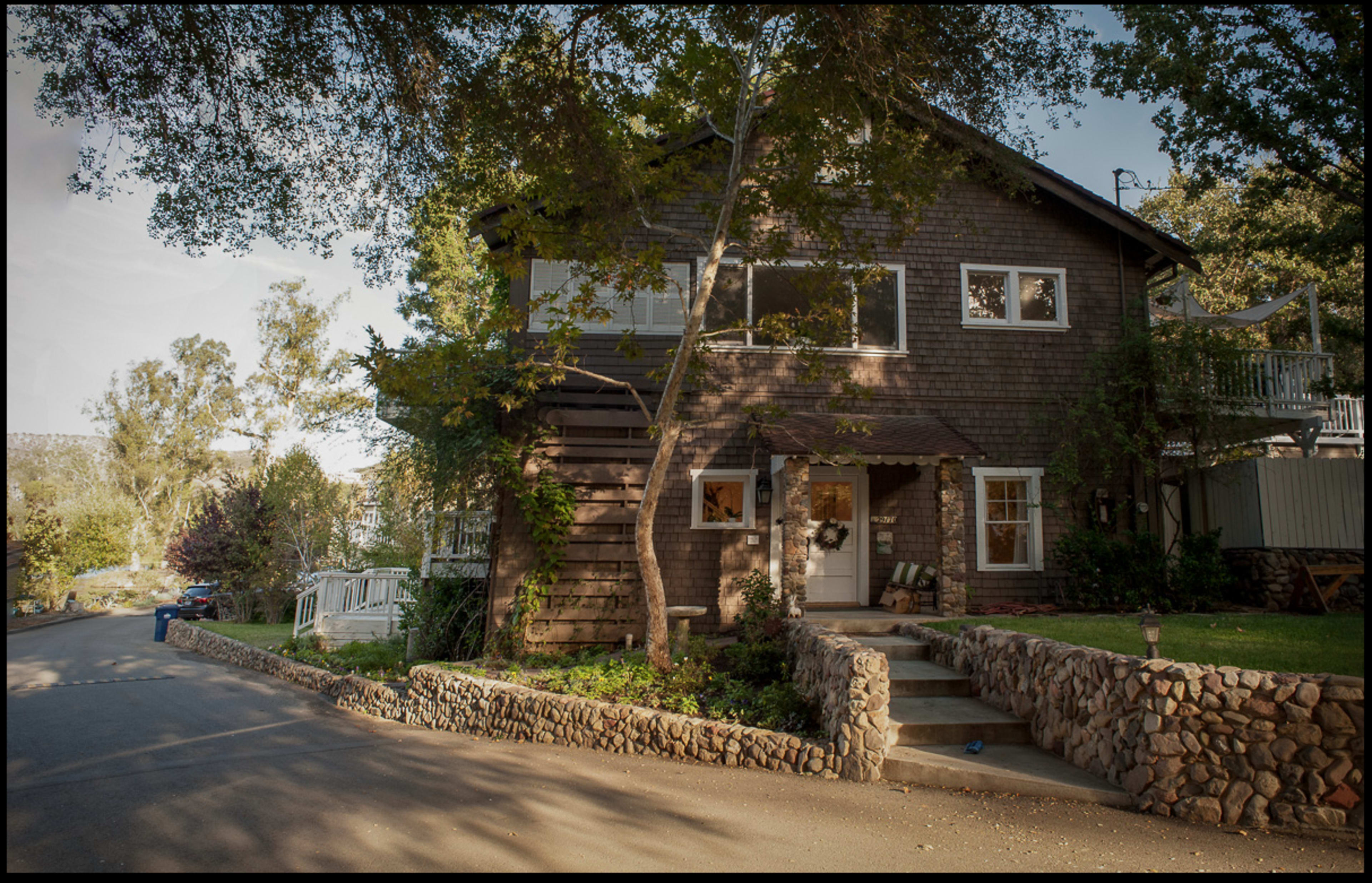 A two-story wooden house with a sloped roof and a stone-bordered garden sits along a tree-lined street.